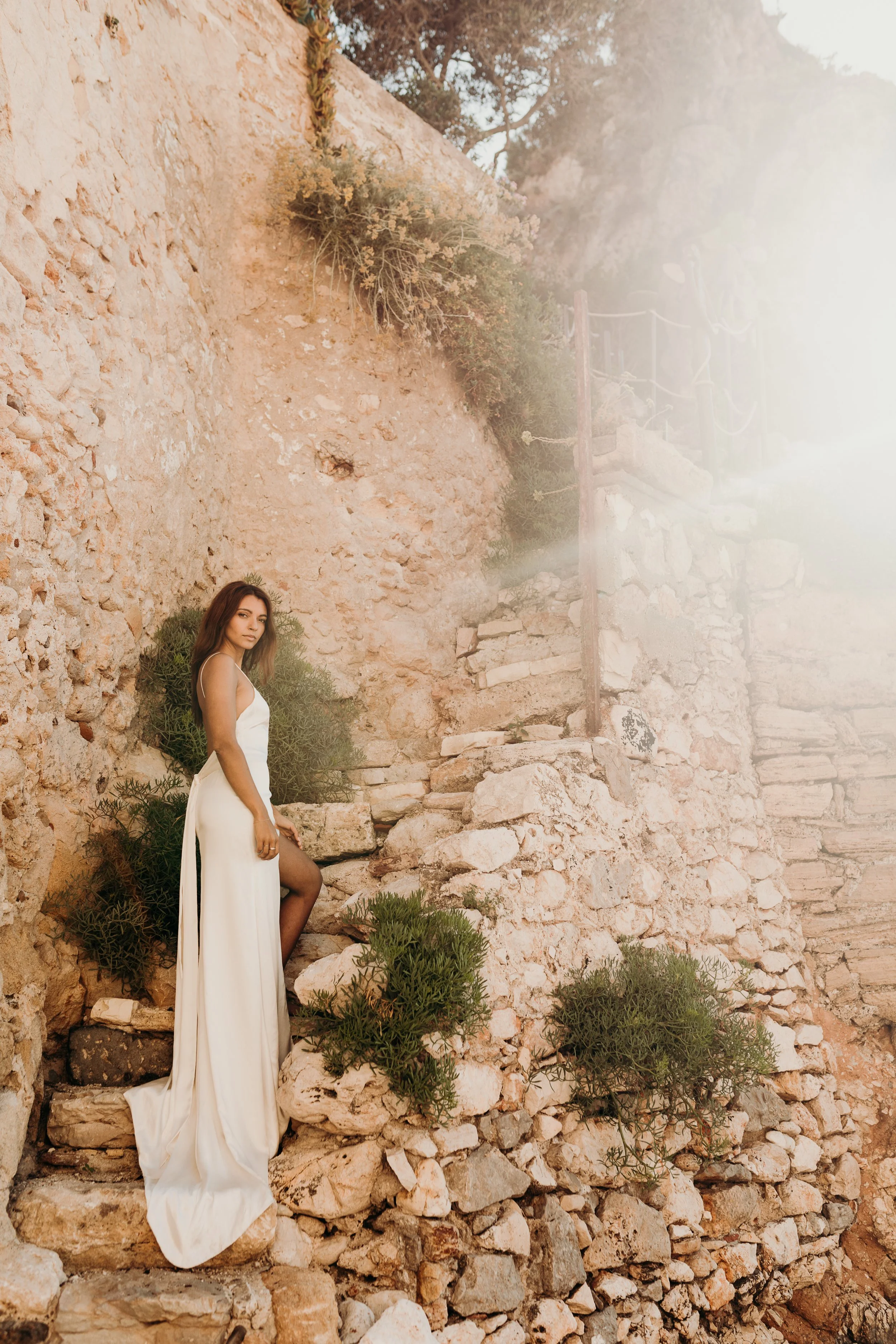 A woman in a white dress standing on rocky steps against a stone wall with plants, and a staircase in the background.