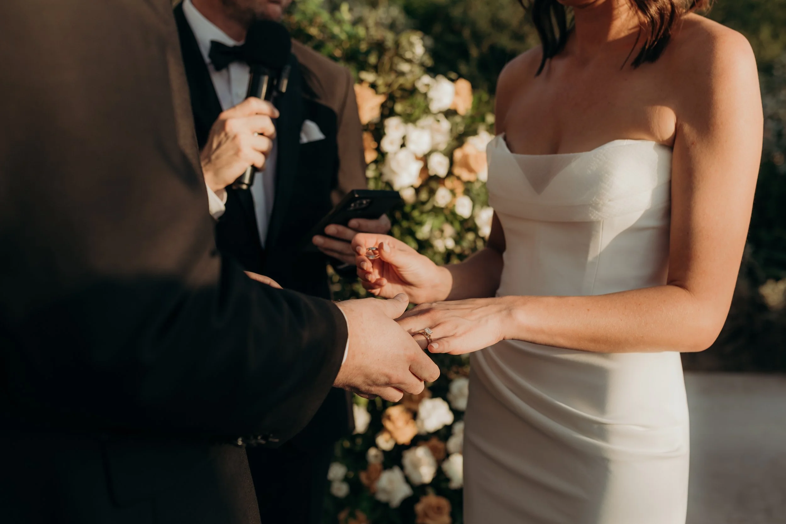 A couple is exchanging wedding rings during a wedding ceremony outdoors, with a man in a tuxedo holding a microphone and a woman in a white strapless wedding dress.