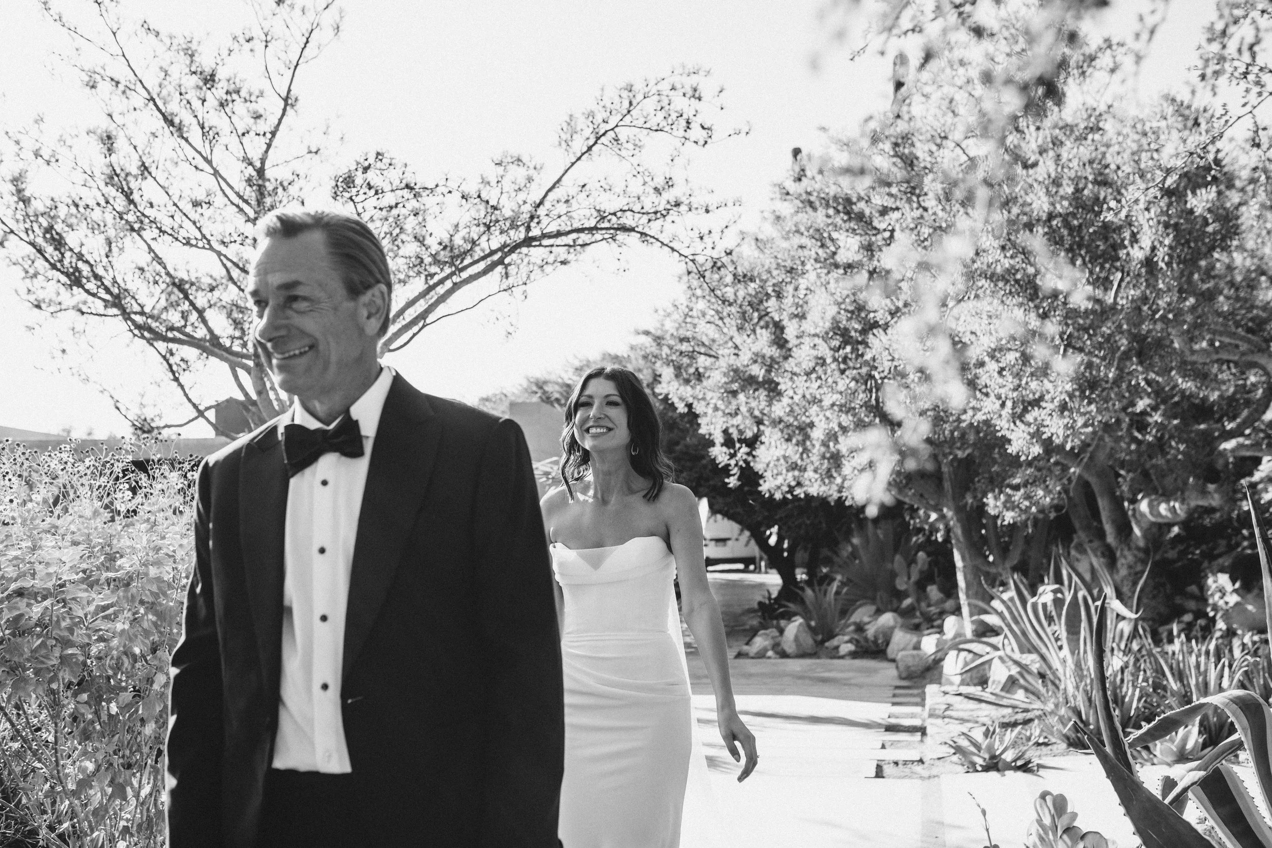 A black and white photo of a smiling older man in a tuxedo and a young woman in a strapless gown walking outdoors among trees and plants.