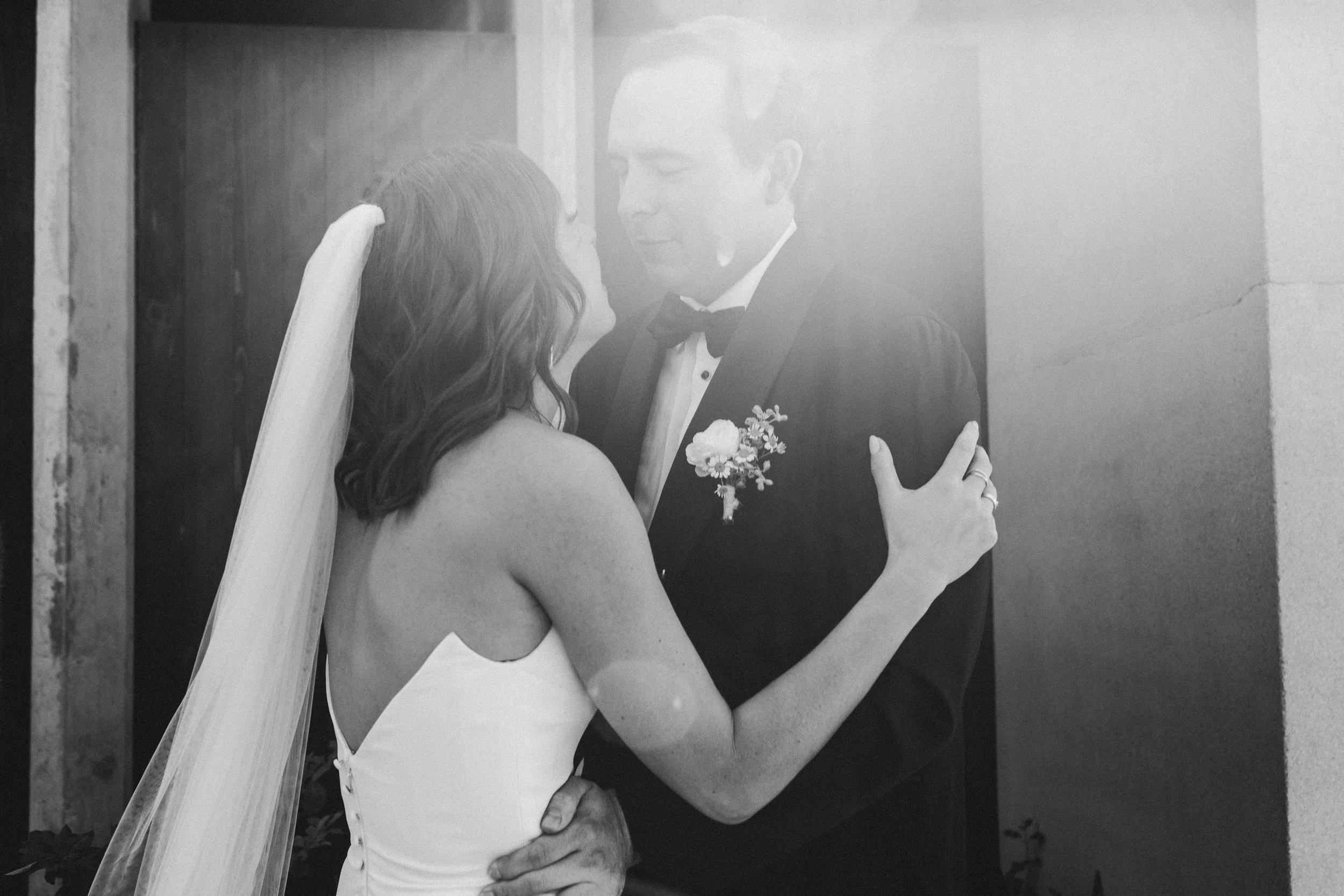 Black and white photo of a bride and groom close to each other, with the bride touching the groom's face and the groom gently holding the bride's waist. They are dressed in wedding attire, and the groom is wearing a tuxedo with a boutonniere.
