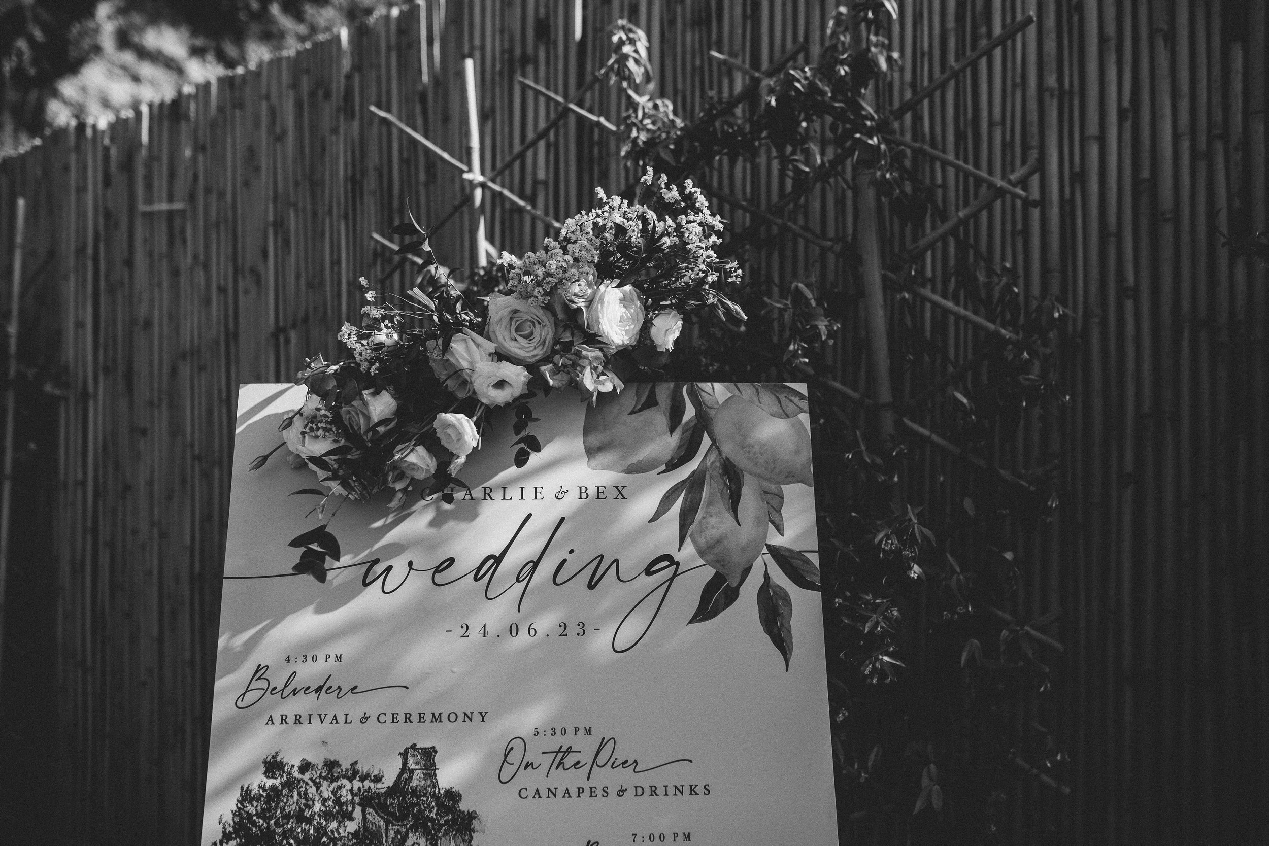 Wedding program with a floral design, listing events such as arrival, ceremony, reception, and drinks, with a background of a bamboo fence and natural scenery.