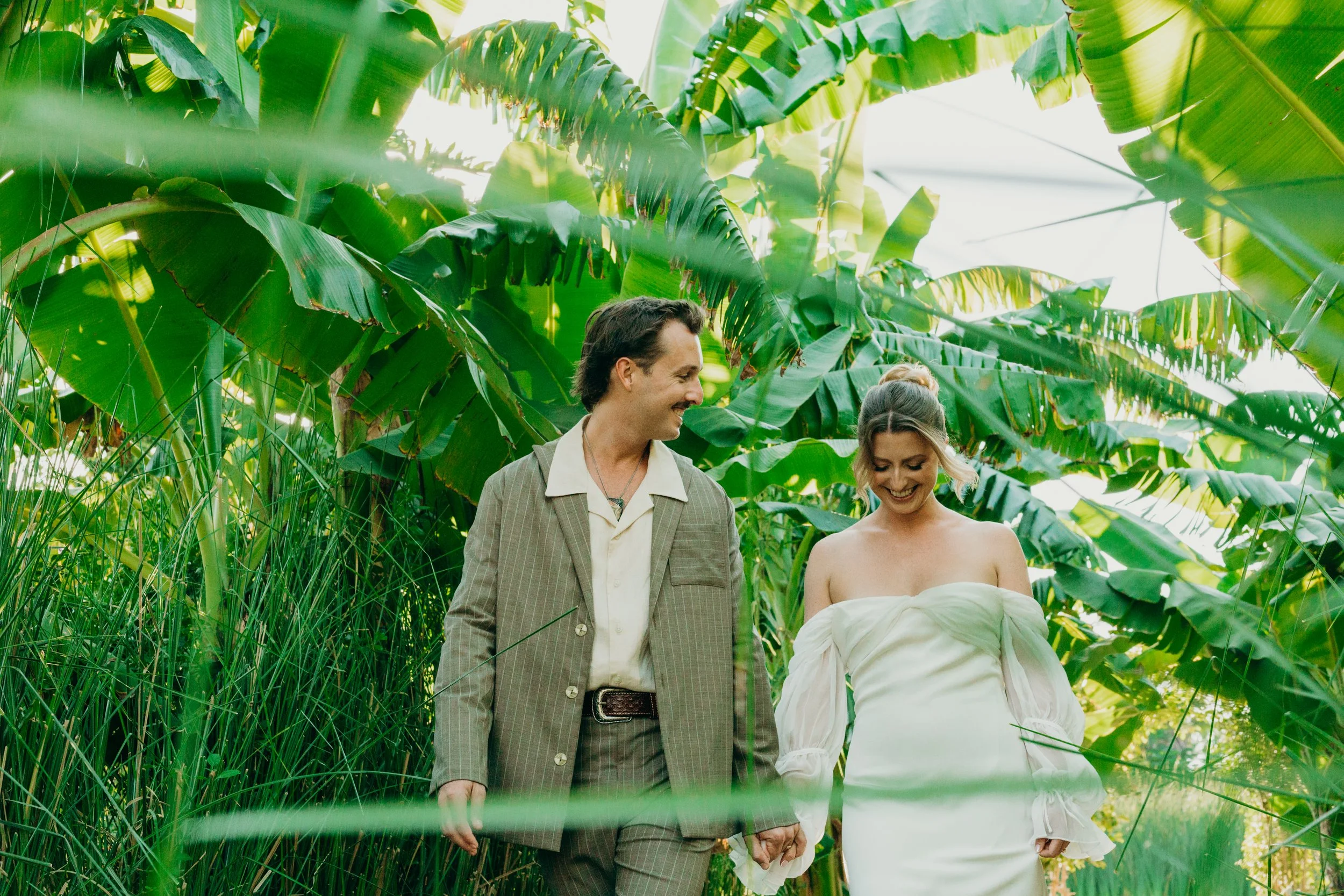 A man and a woman are holding hands and walking through a lush green jungle with large leaves and tall grass, smiling and looking at each other.