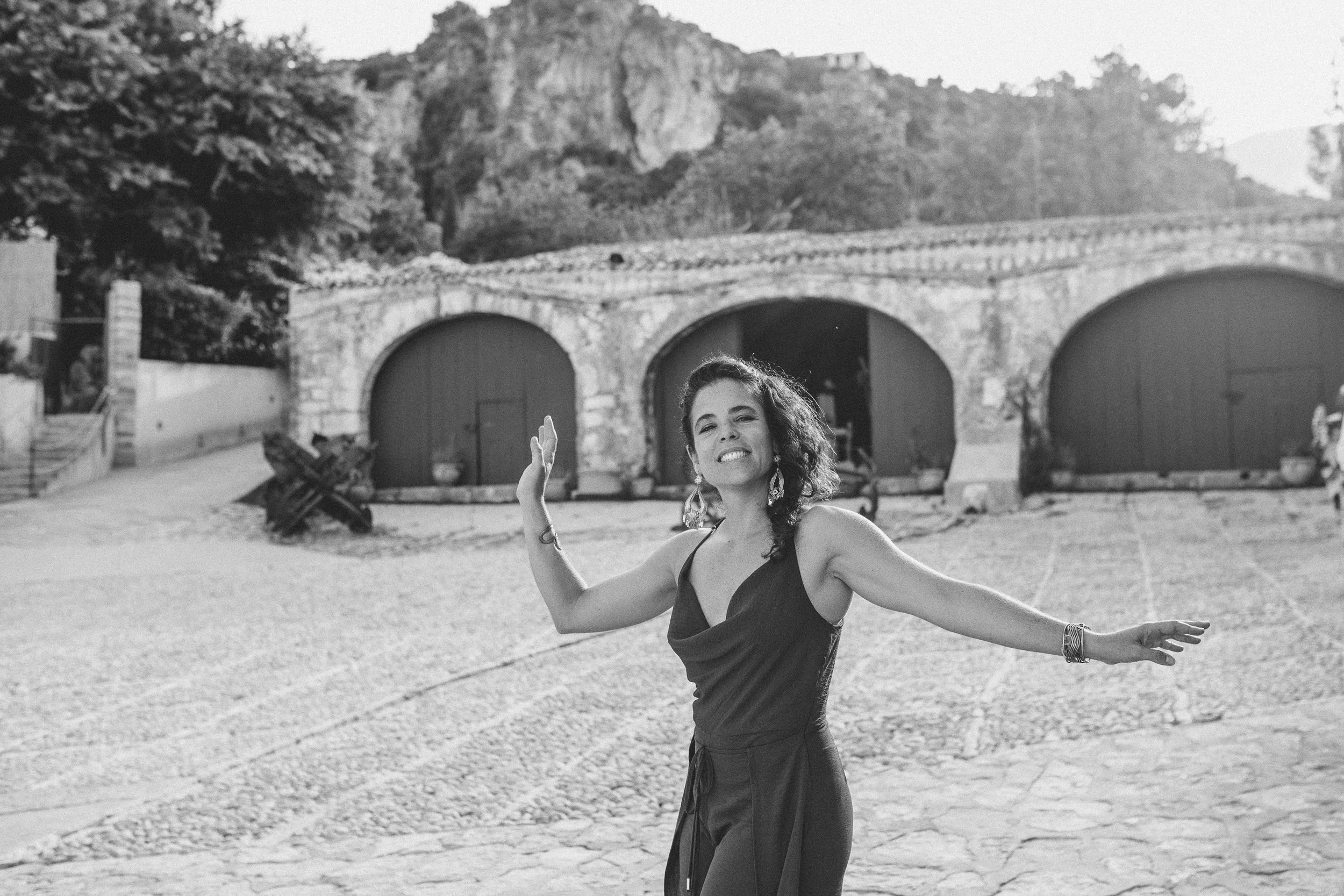 A smiling woman with curly hair, wearing a sleeveless dress and jewelry, dancing outdoors on a sunny day, with stone buildings and a hill in the background.