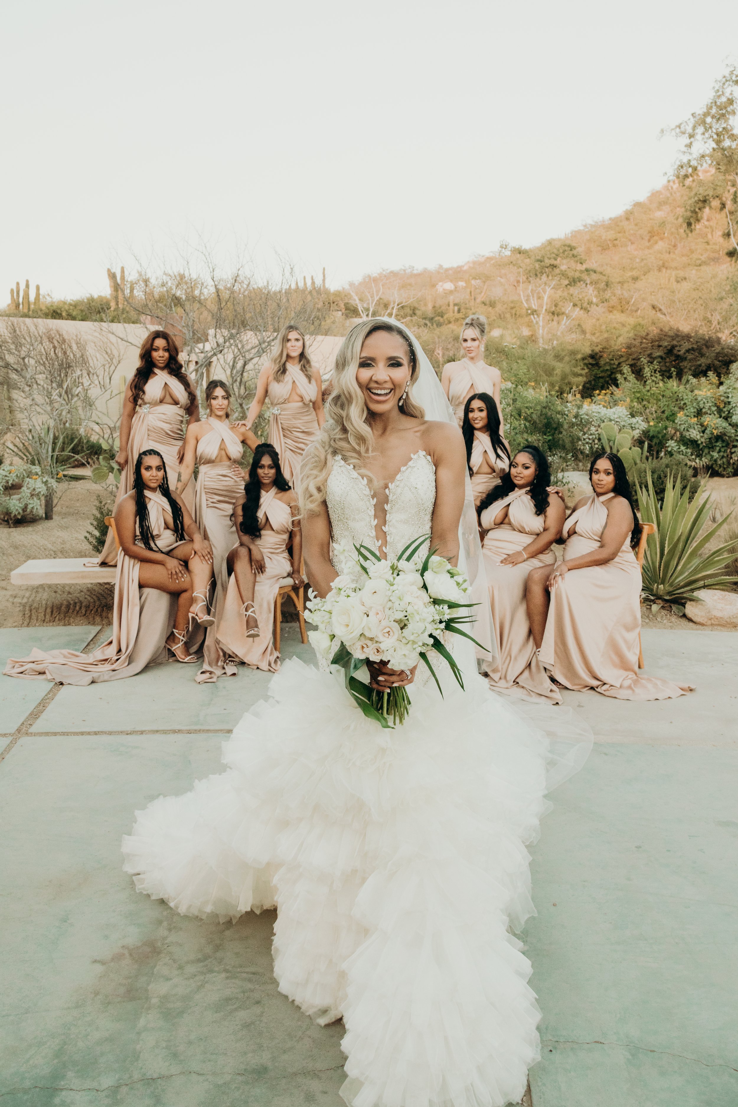 A bride in a white wedding gown holding a bouquet of white flowers, smiling, with ten bridesmaids in beige dresses with cutouts and draped necklines in the background, outdoors with desert plants and hills in the background.