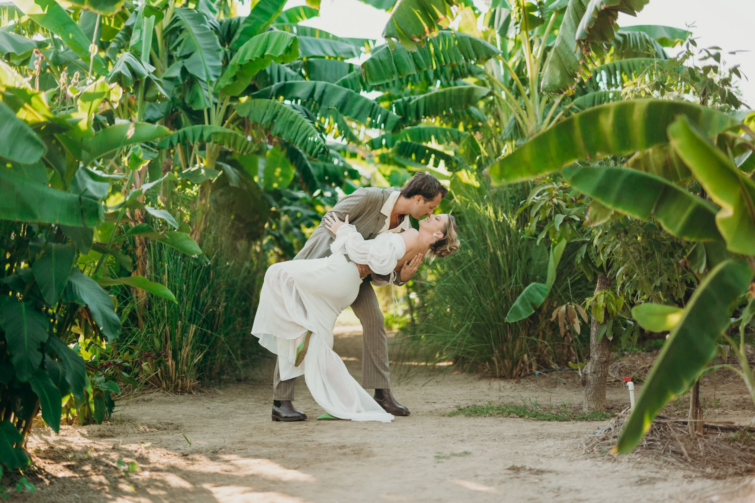 A man in vintage clothing is dipping a woman in a white dress, with a lush green garden filled with large leafy plants in the background.