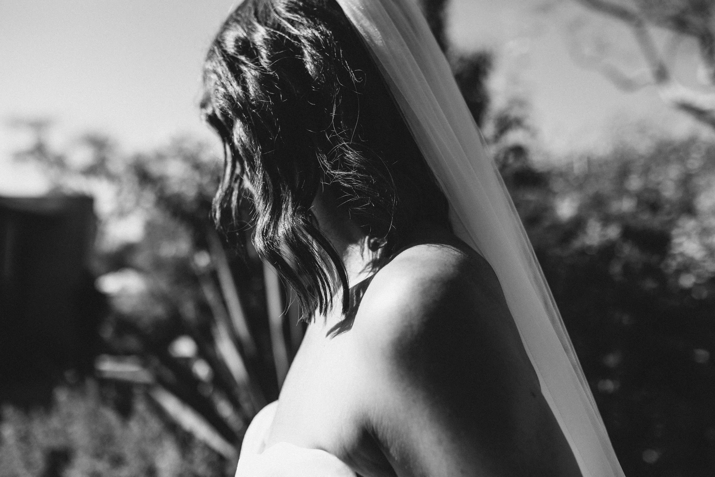 A black and white photo of a person with wavy hair holding a surfboard on their shoulder, standing outdoors with blurred trees and sky in the background.