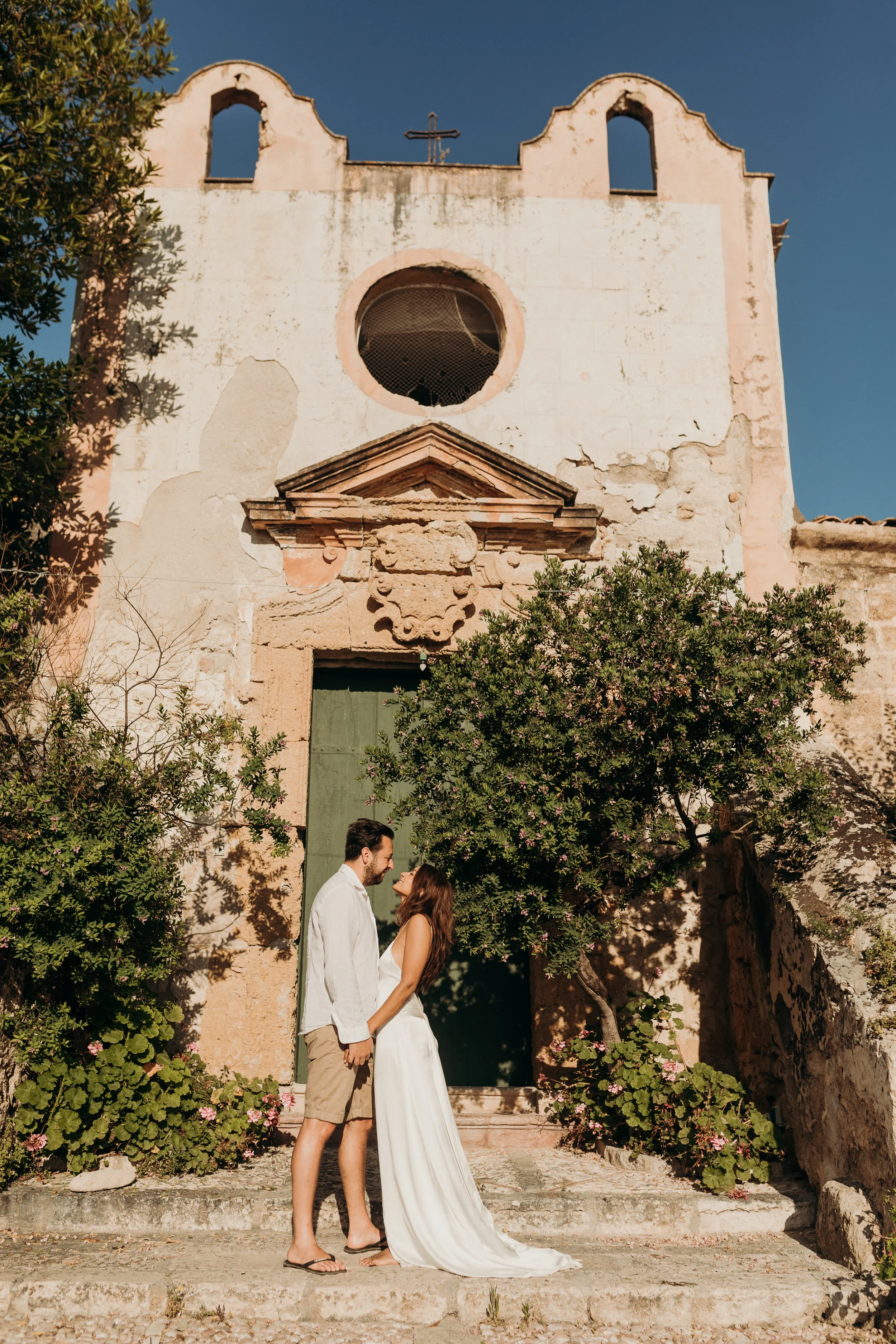 A couple stands close together holding hands outside a weathered historic church with a large circular window and an old moss-covered cross on top. The man wears a white shirt and beige shorts, and the woman wears a white dress. They are surrounded b