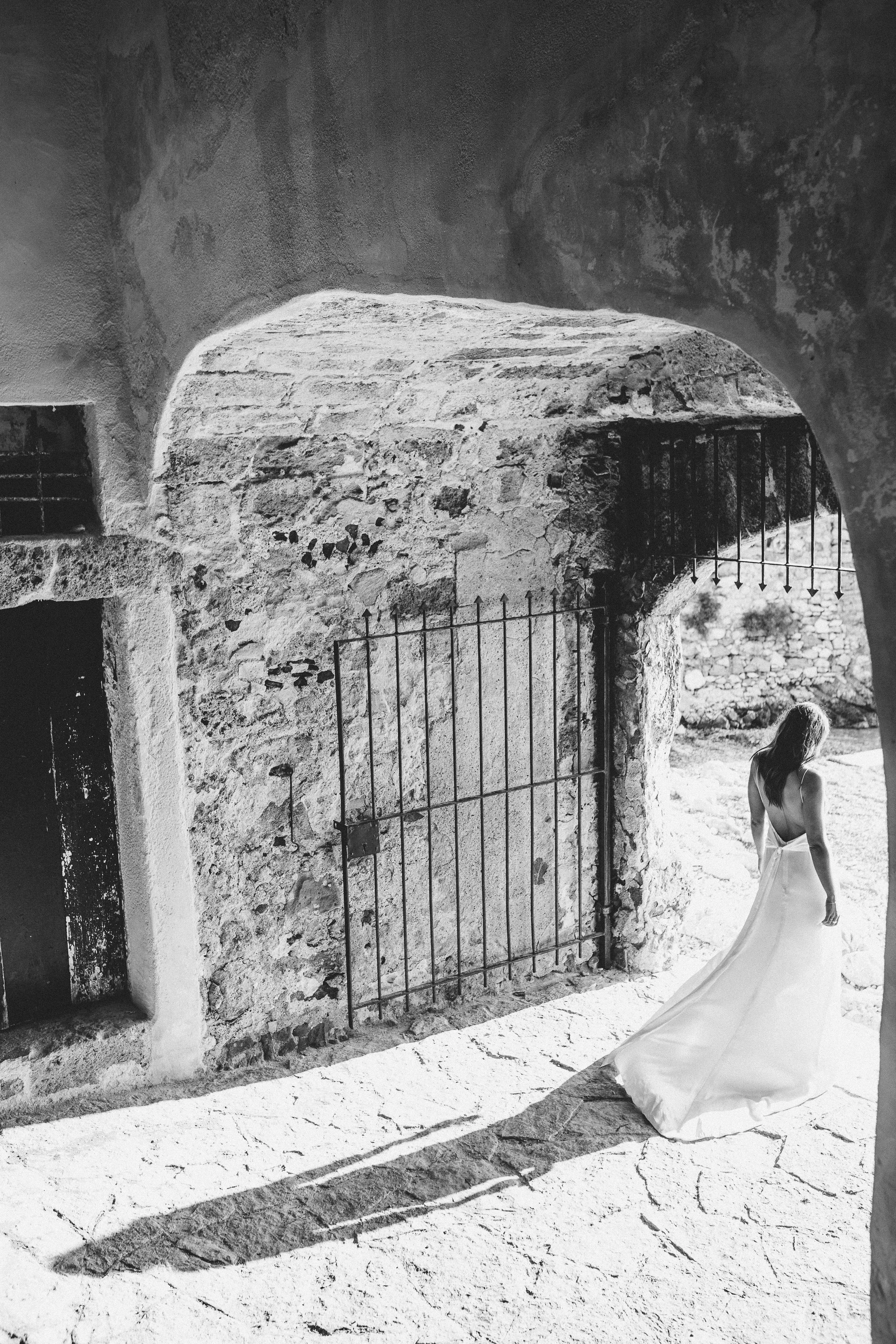 A woman in a wedding dress walking through an arched stone doorway with a wrought iron gate, casting a long shadow on a cracked stone floor.