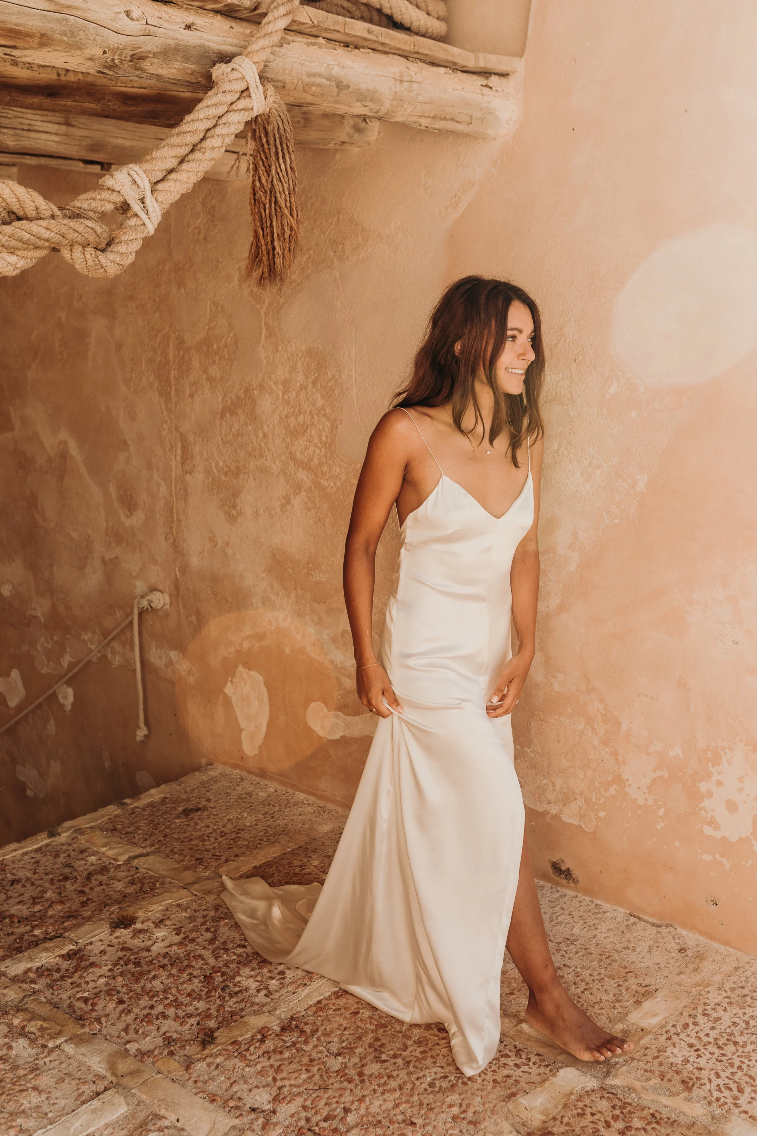 A woman in a white wedding dress standing against a beige textured wall with a rustic wooden and rope decor element.