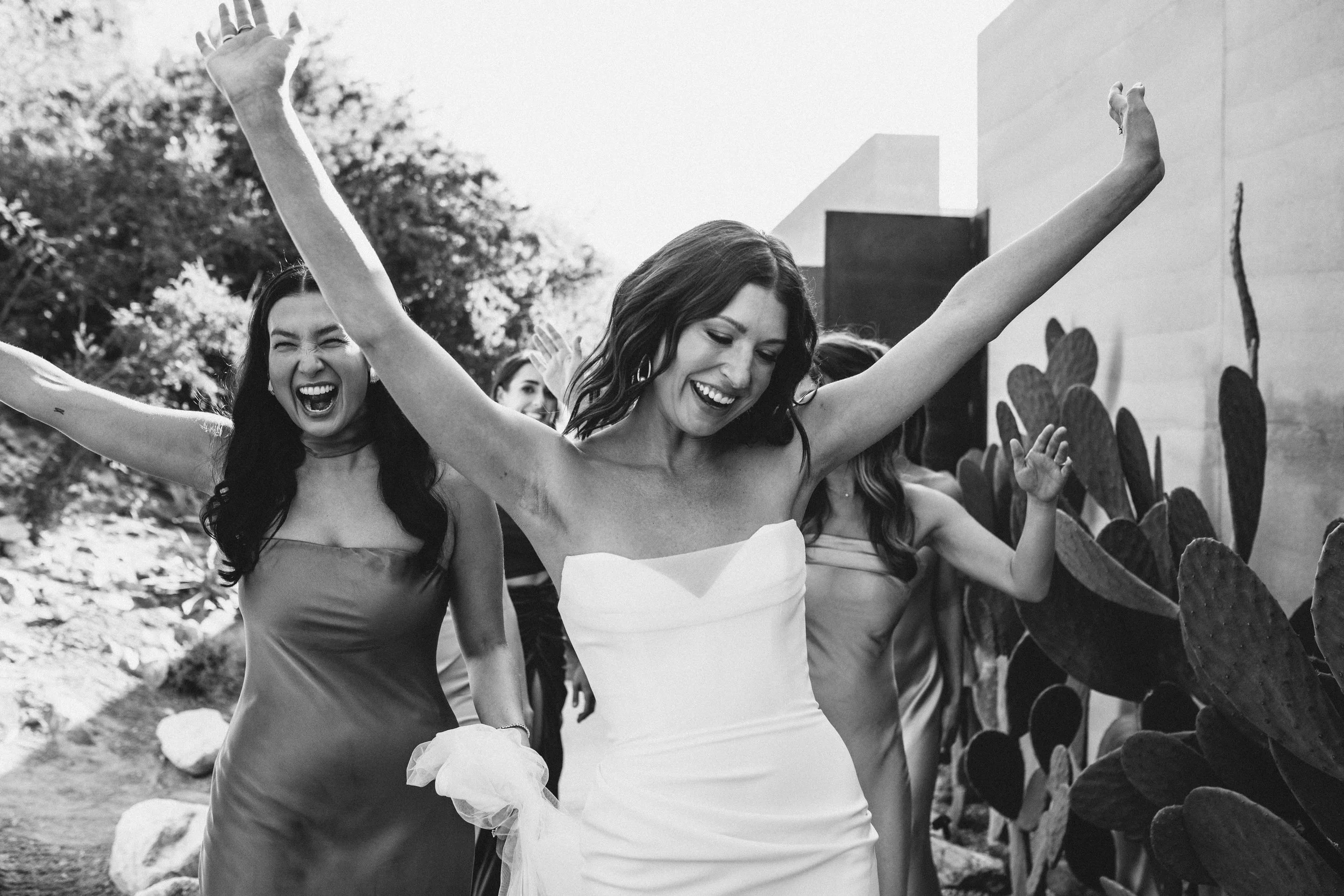 Black and white photo of three women celebrating. They are smiling, with one woman in a strapless dress and others in dresses. They have their arms raised, and one woman is wearing a ring. They are outdoors with trees and a wall behind them.