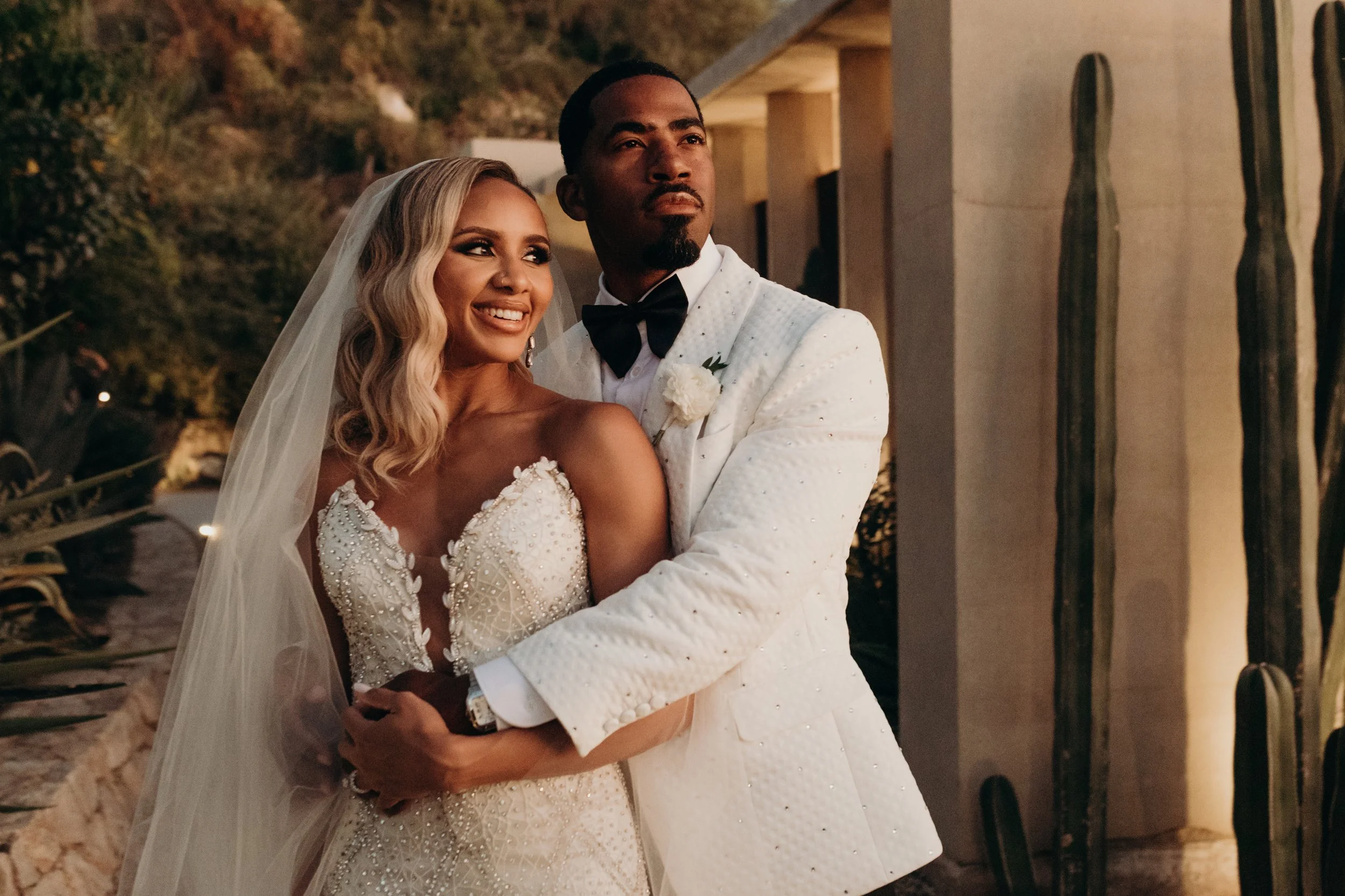 A bride and groom are posing for a photo during their wedding at dusk. The bride has long blonde curly hair and is wearing a white wedding dress with intricate beading, and the groom has short dark hair and a goatee, dressed in a white tuxedo with a 