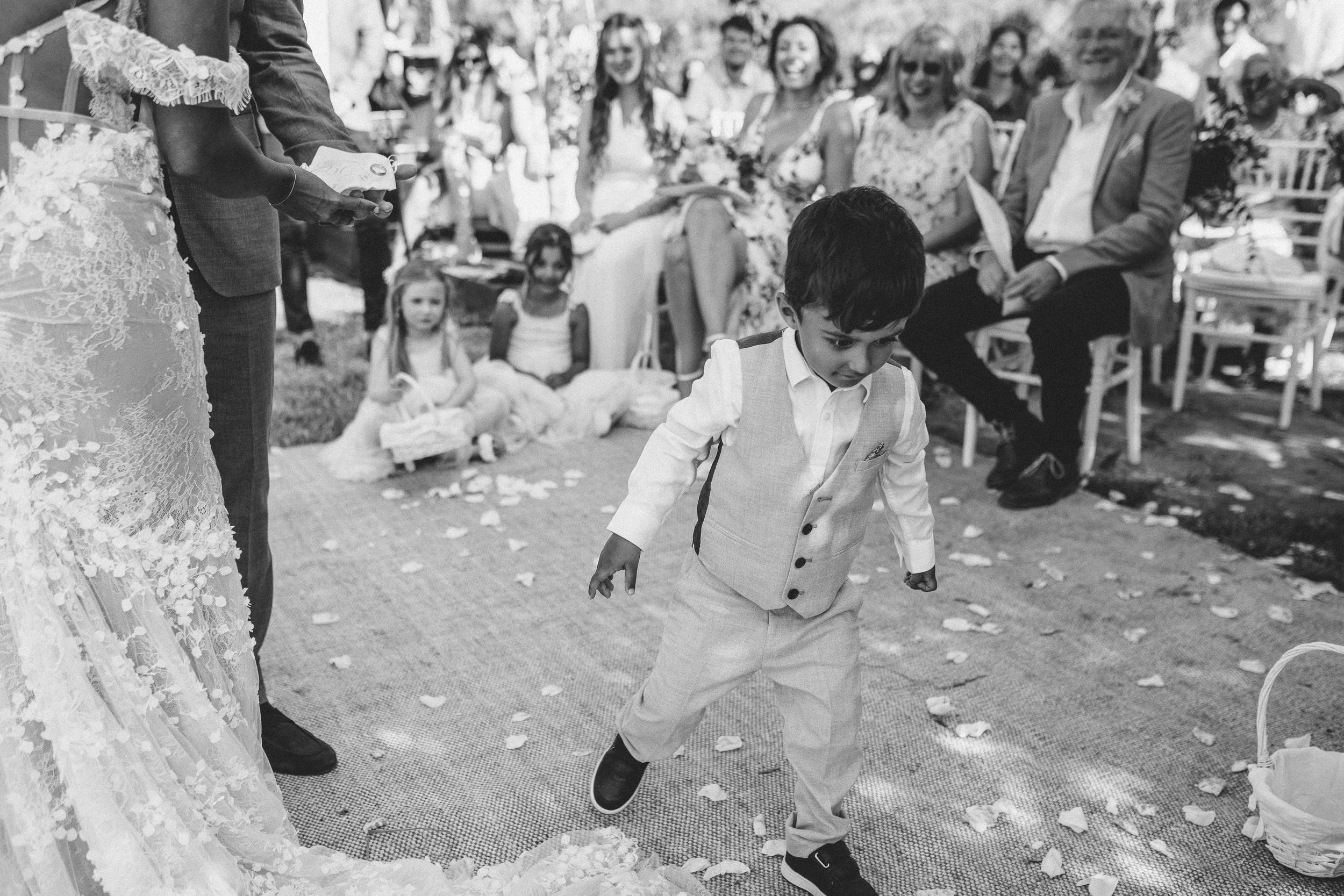 Children participating in a wedding rice-throwing game outdoors, with adults and women in floral dresses observing in the background, flowers and chairs set up for the event.