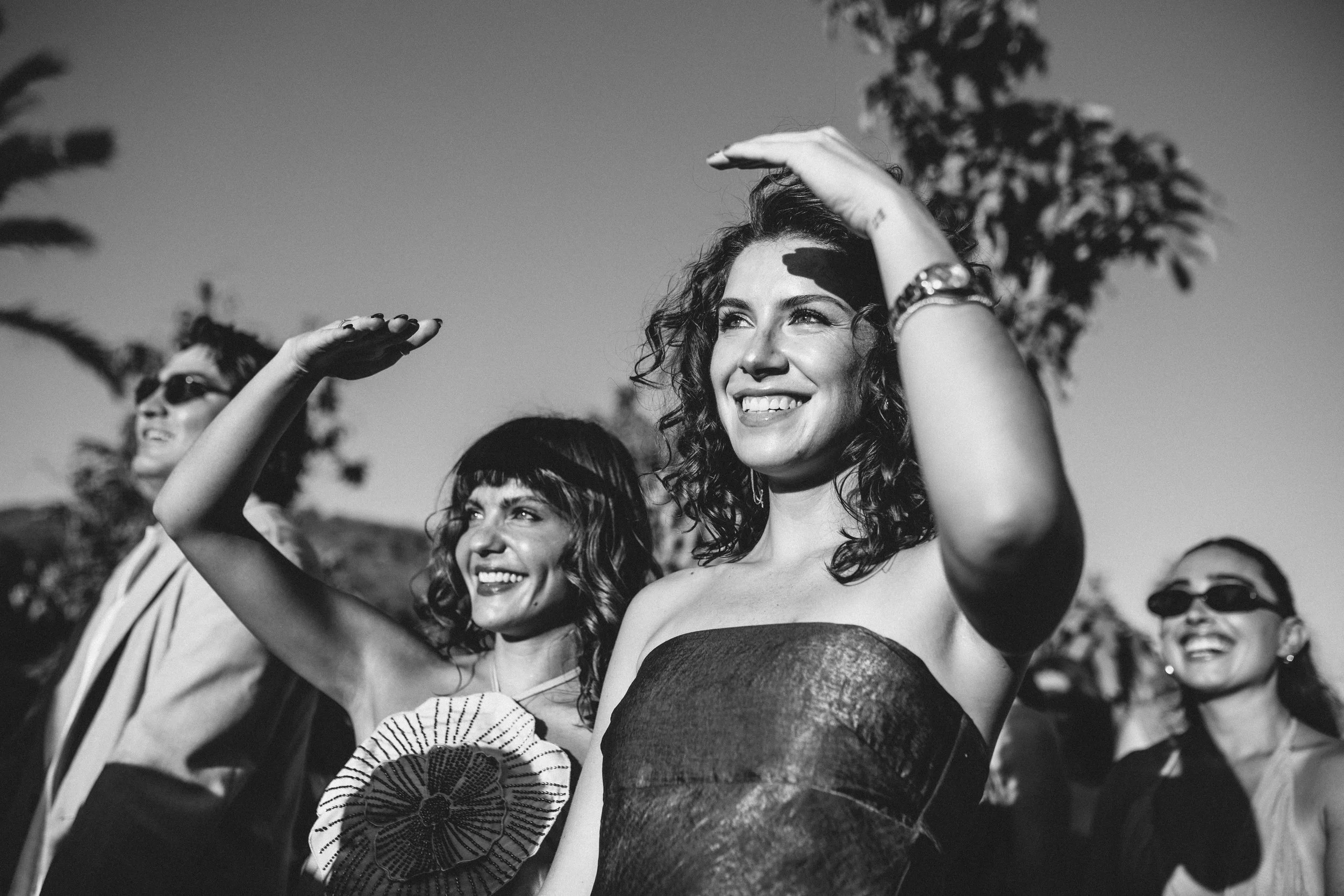 Black and white photo of women smiling and waving outdoors during daylight. The woman in the foreground has curly hair and is wearing a strapless dress, while others behind her are wearing sunglasses and clothing with patterns. The background shows t