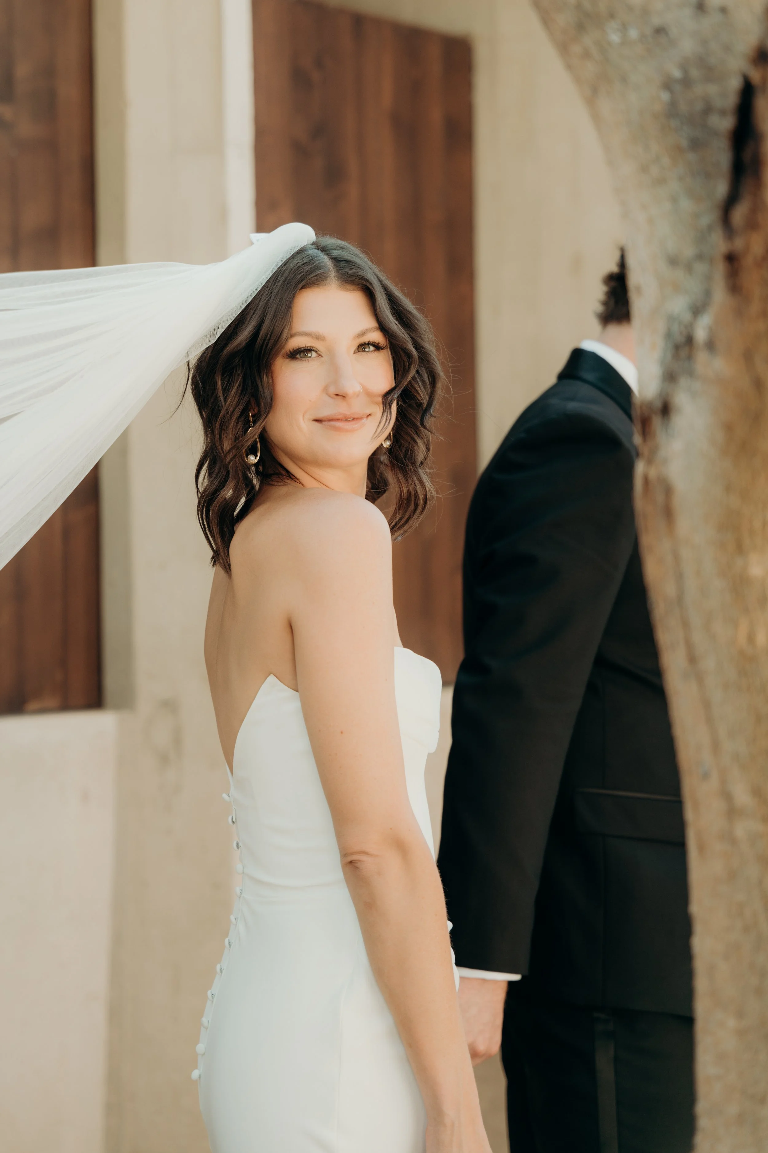 A bride with shoulder-length brown hair and a smile, wearing a white strapless wedding dress with buttons on the back, is standing next to a man in a black tuxedo, who is partially visible, inside a building with wooden and stone walls.
