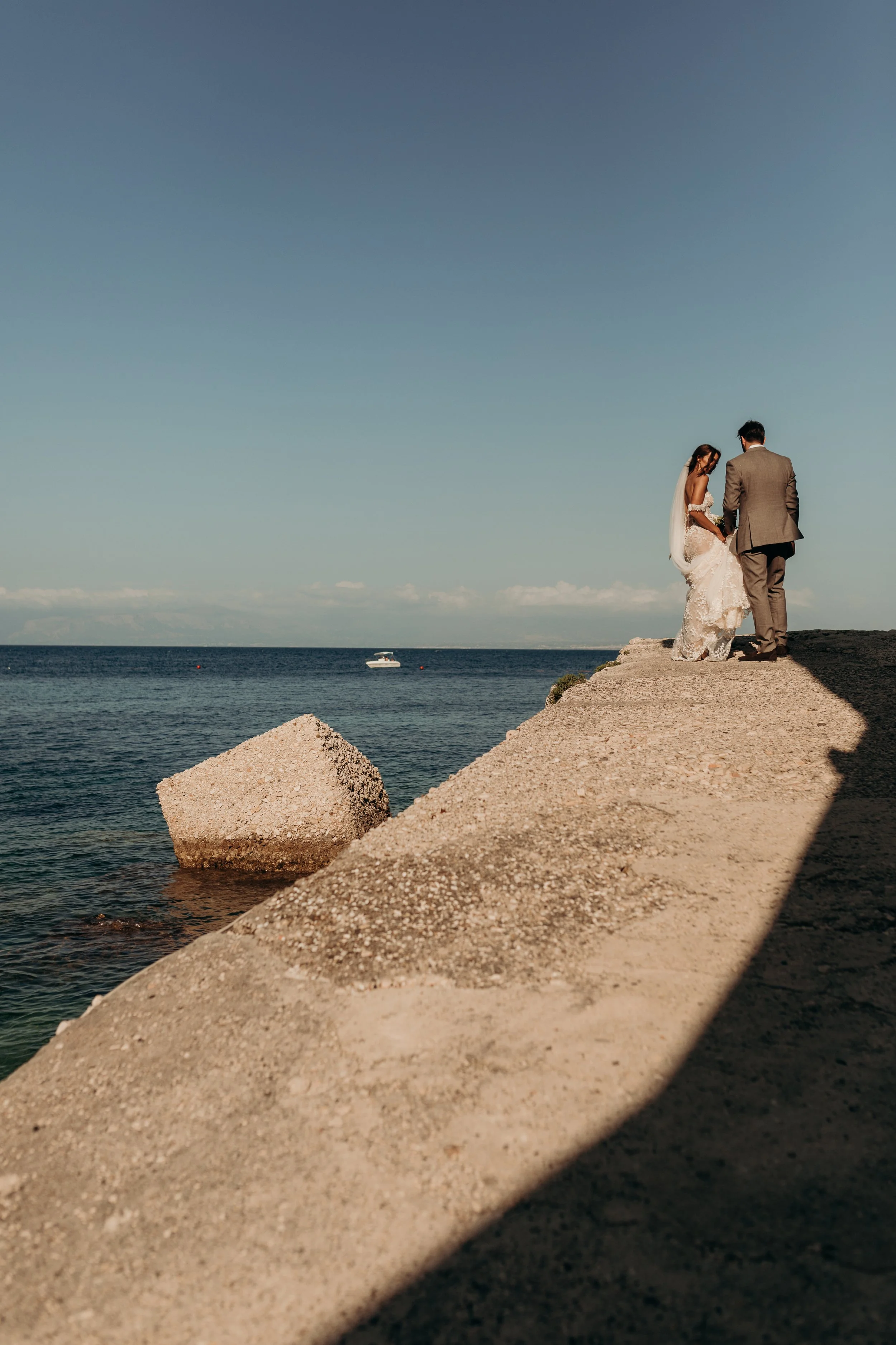 A bride and groom standing on a concrete pier by the ocean, holding hands and looking at each other, with a boat in the water and a clear blue sky.