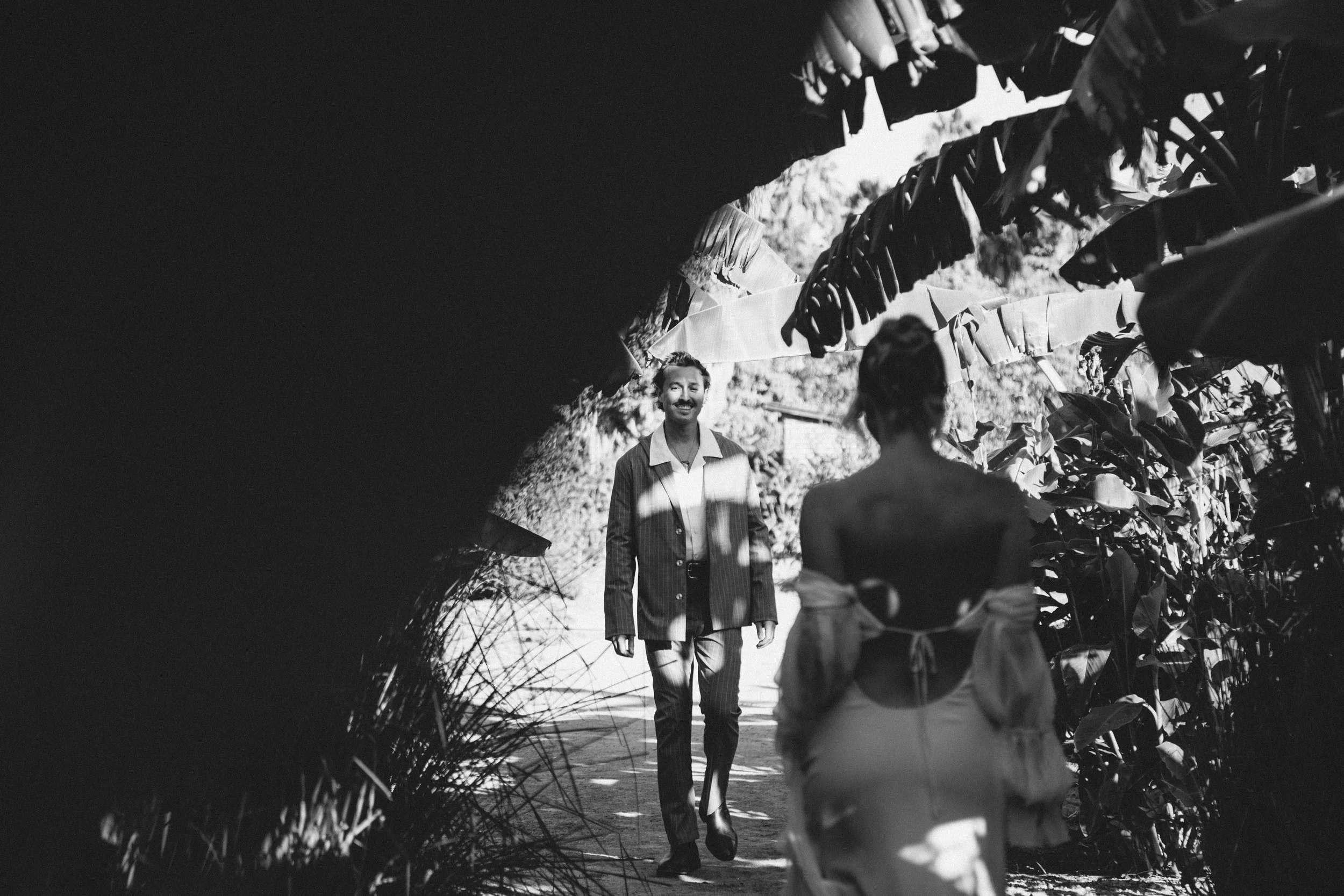 Black and white photo of two people walking outdoors, surrounded by leafy plants and trees. One person is in the foreground, the other is smiling in the background.