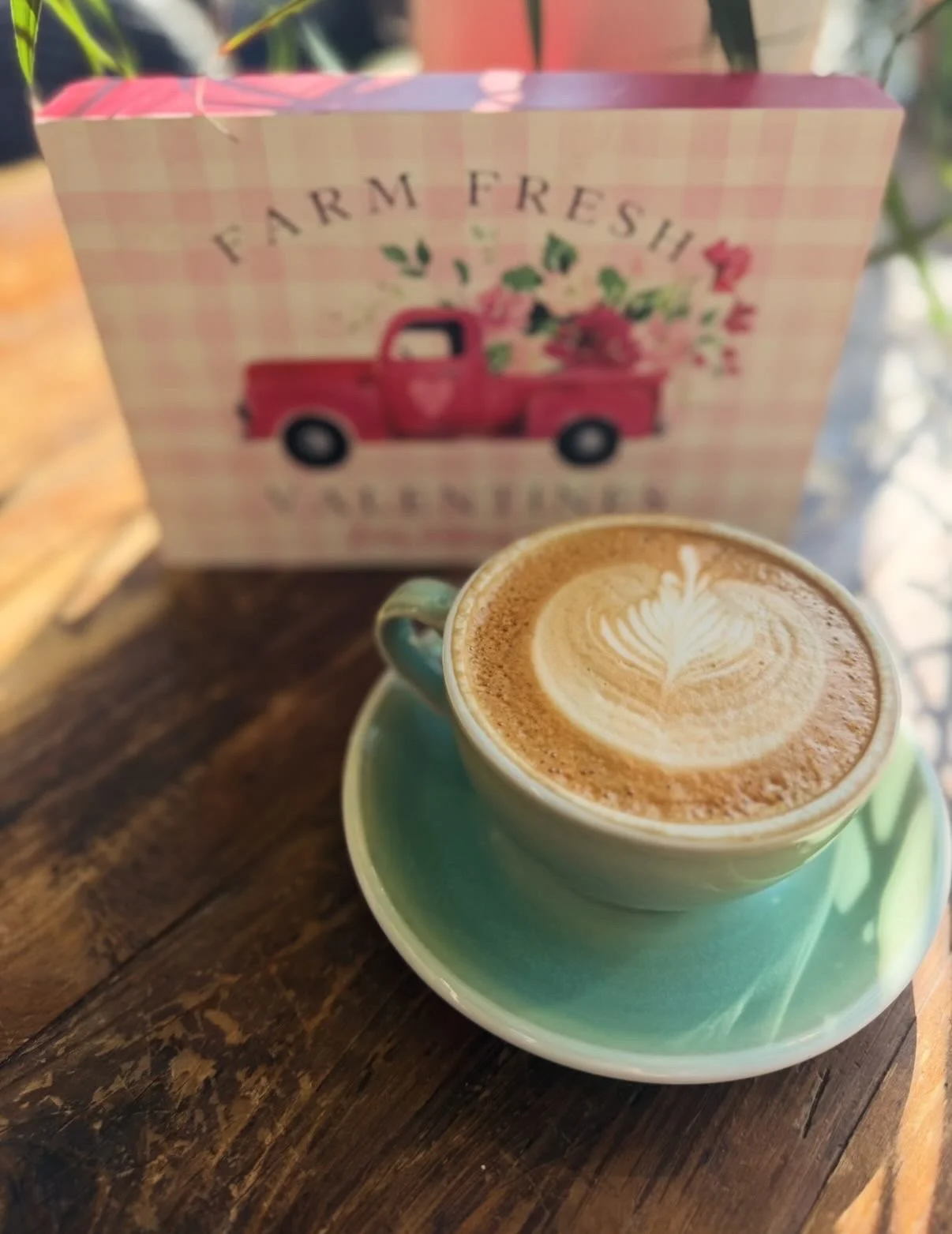 A cup of latte with latte art on top, placed on a turquoise saucer on a wooden table, with a pink checkered box with a vintage red truck and floral design in the background.