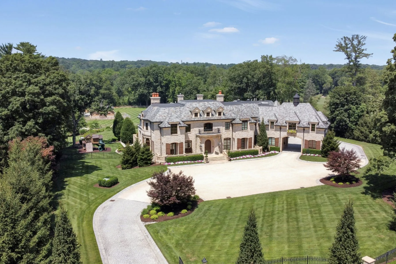 Large estate with a grand stone mansion surrounded by well-manicured lawns, colorful flower beds, tall trees, and a curved driveway in a rural landscape.