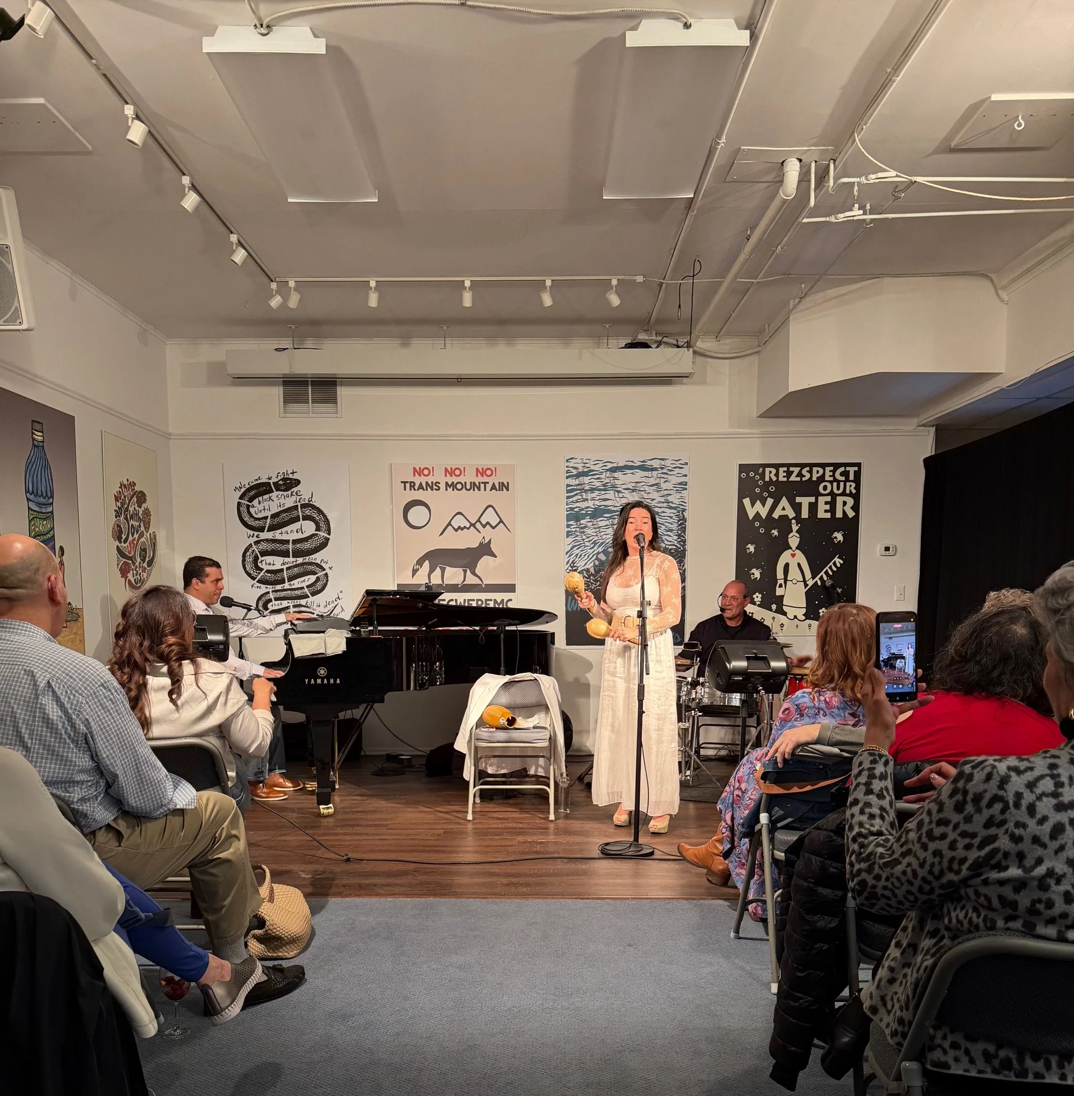 A woman in a white dress singing into a microphone on a stage with a band, including a pianist, a drummer, and an audience watching.