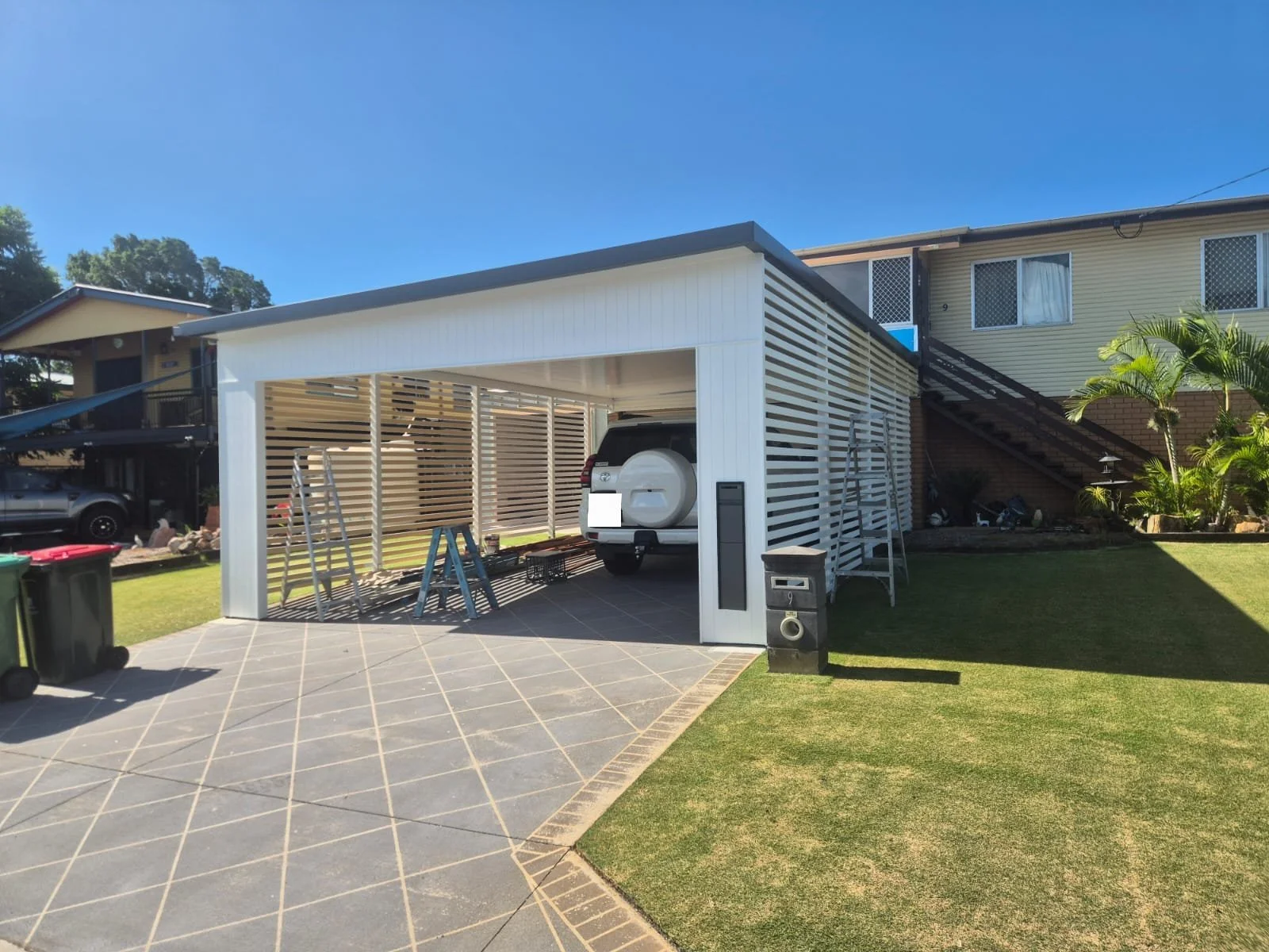 Carport under construction with a white wooden frame, ladder, tool boxes, and a white SUV inside, next to a yellow house with outdoor stairs and green plants.