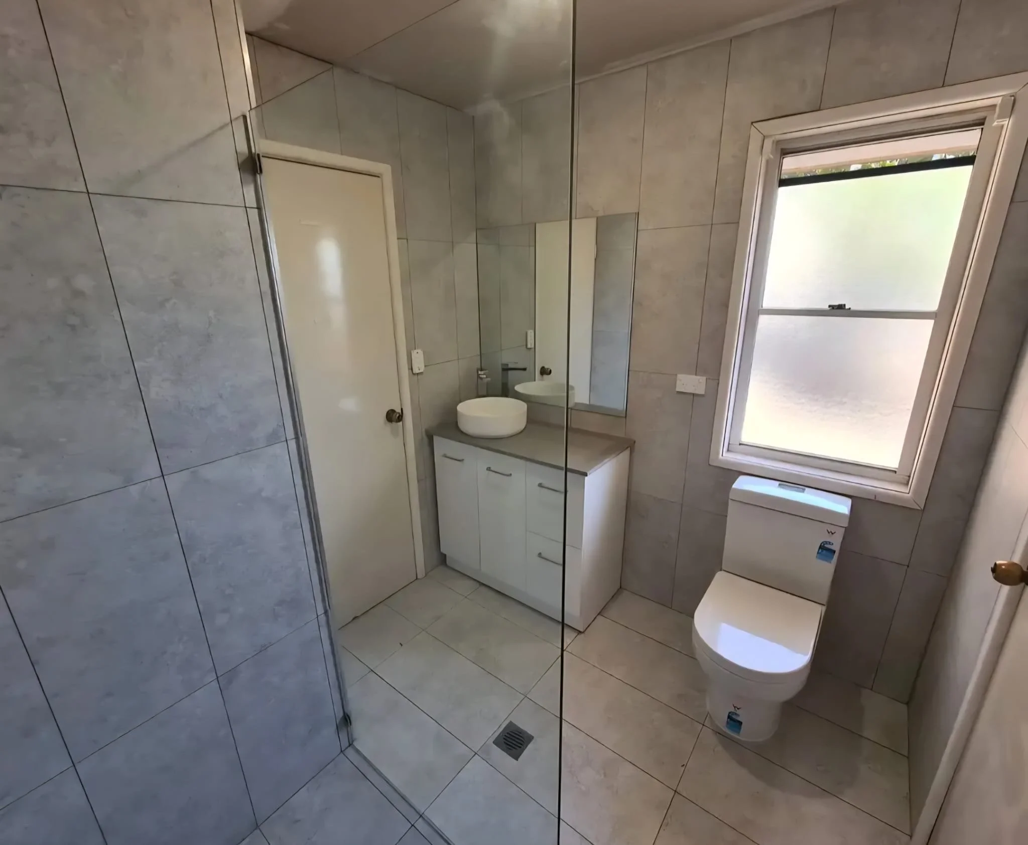 A clean bathroom with gray tile walls and floor, a white toilet next to a window, a white vanity with a vessel sink, and a large mirror above the sink.