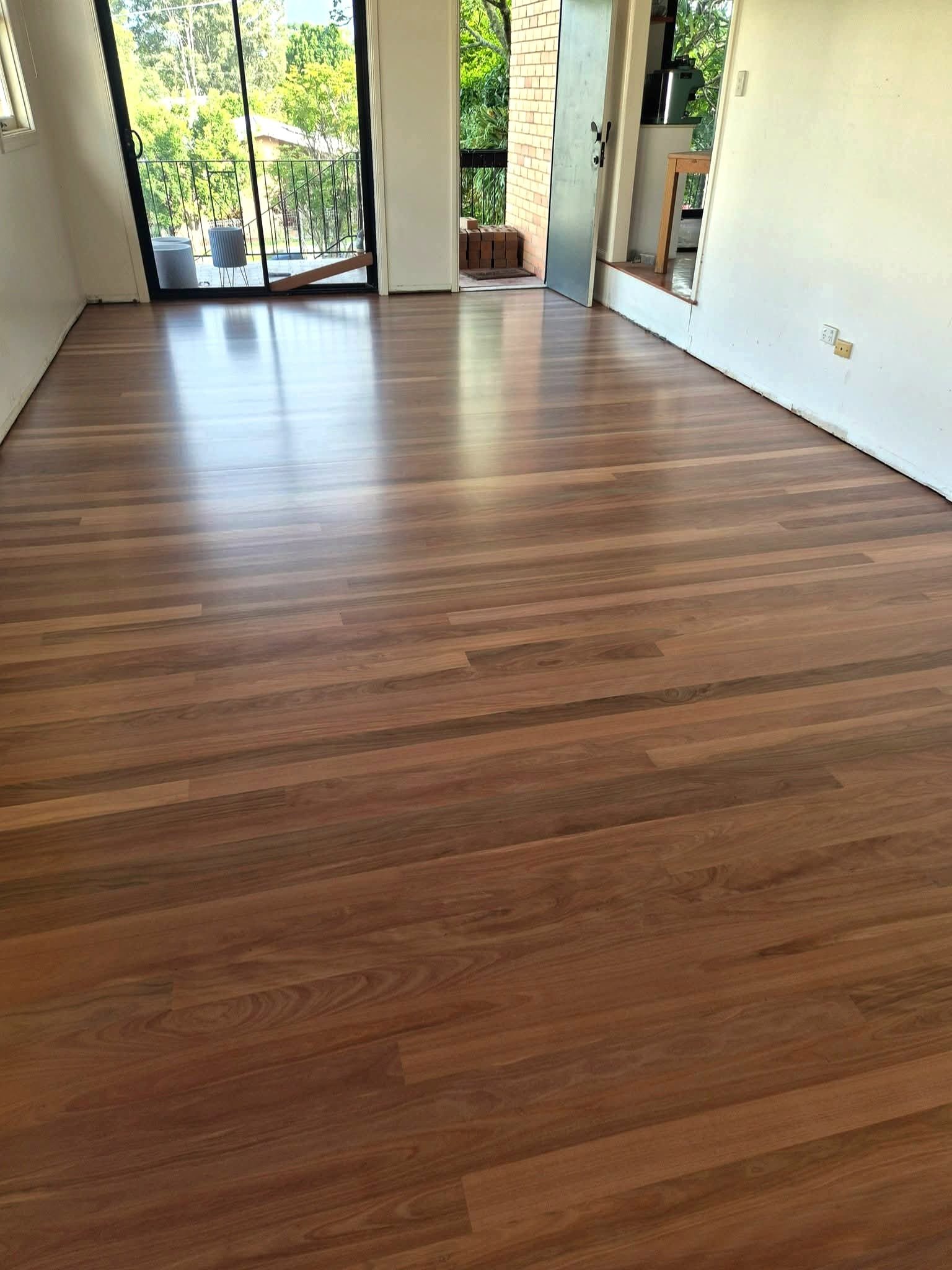 An empty room with wooden flooring, a sliding glass door leading to a balcony, and a small section of a kitchen visible through a doorway.