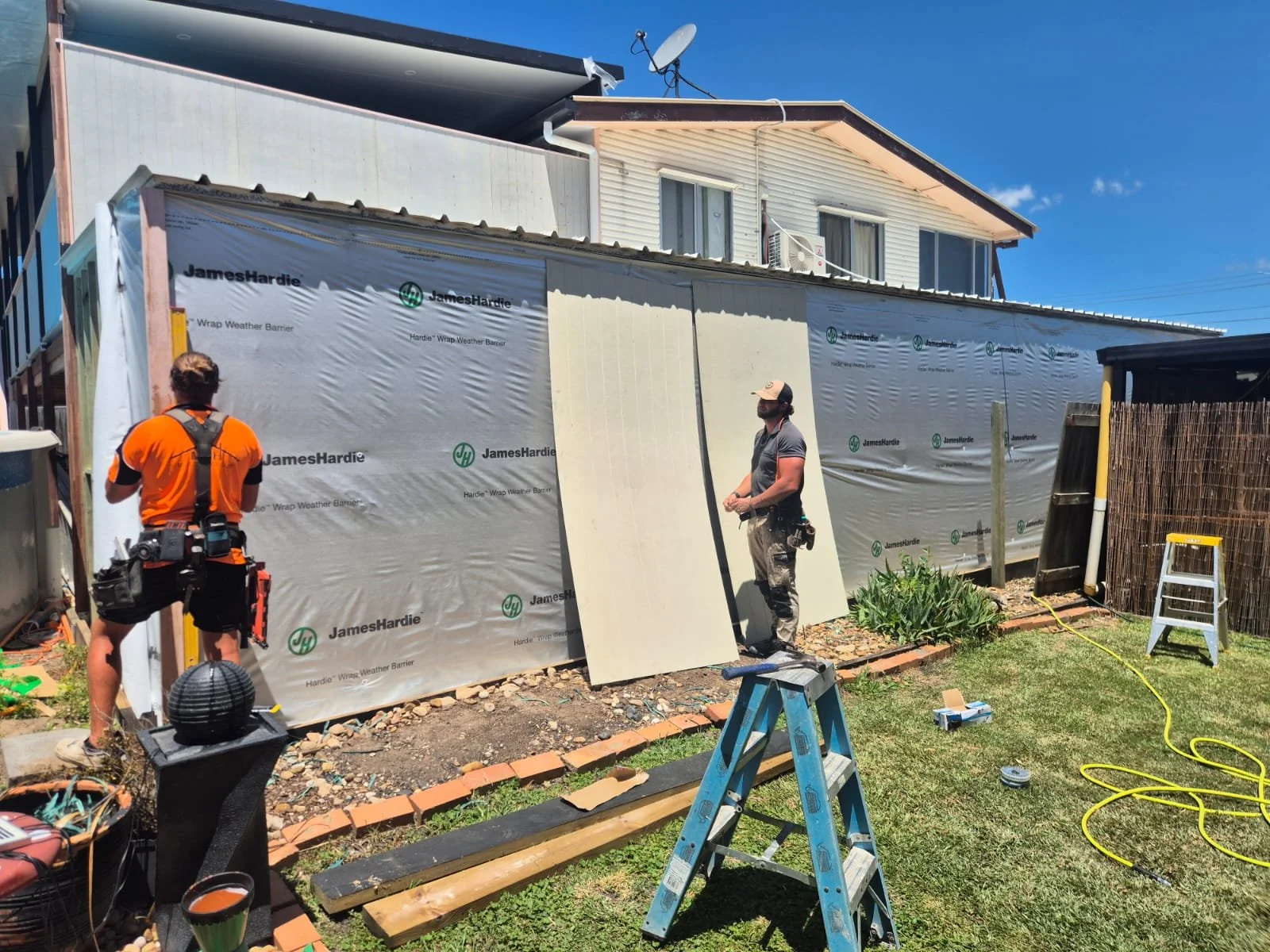 Construction workers install weatherproofing on the side of a house with JamesHardie branded wrap, outside in a sunny yard with tools, ladder, and bricks.