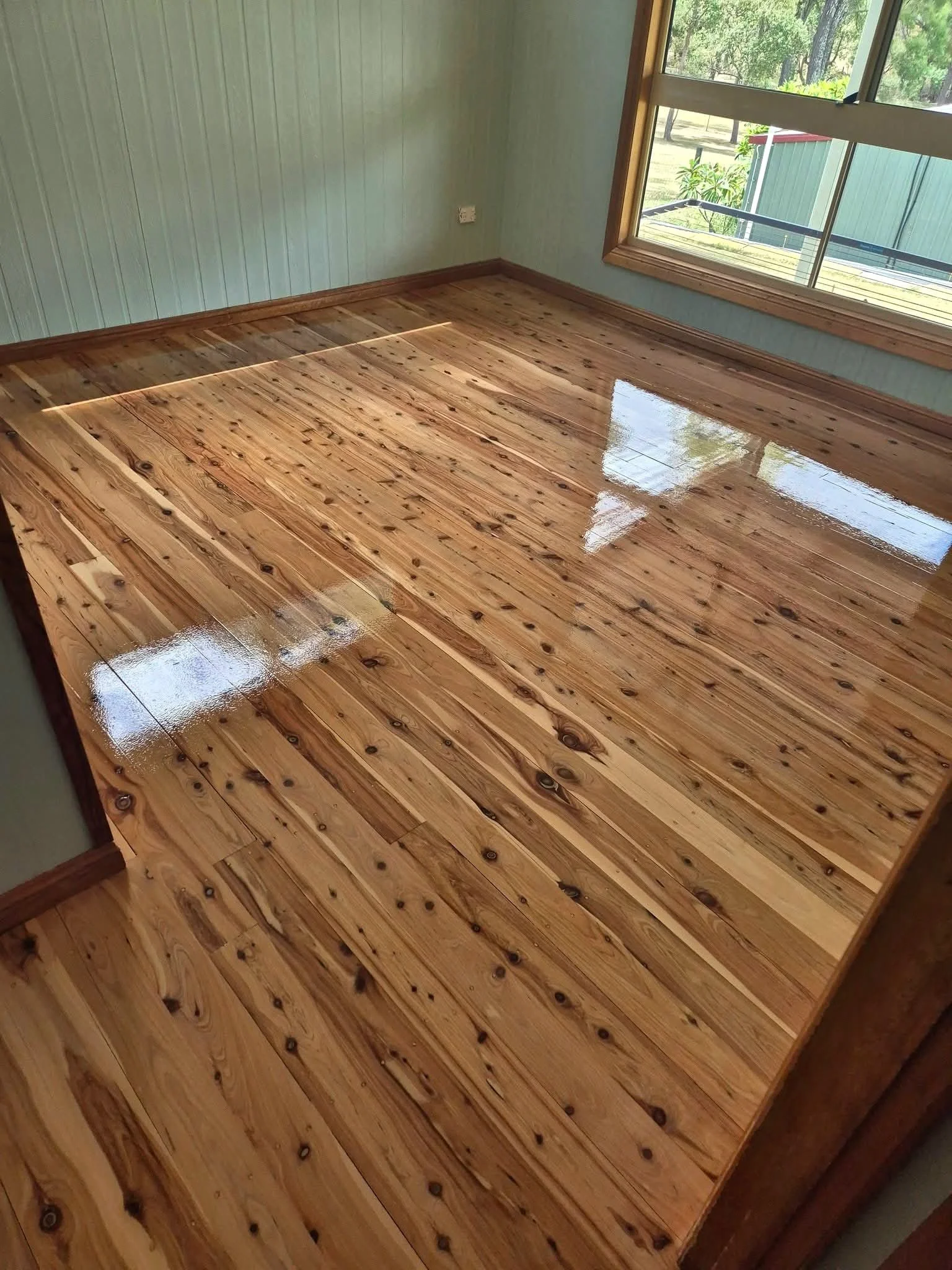 A room with freshly polished wooden floorboards, light green walls, and large window showing outdoor greenery.