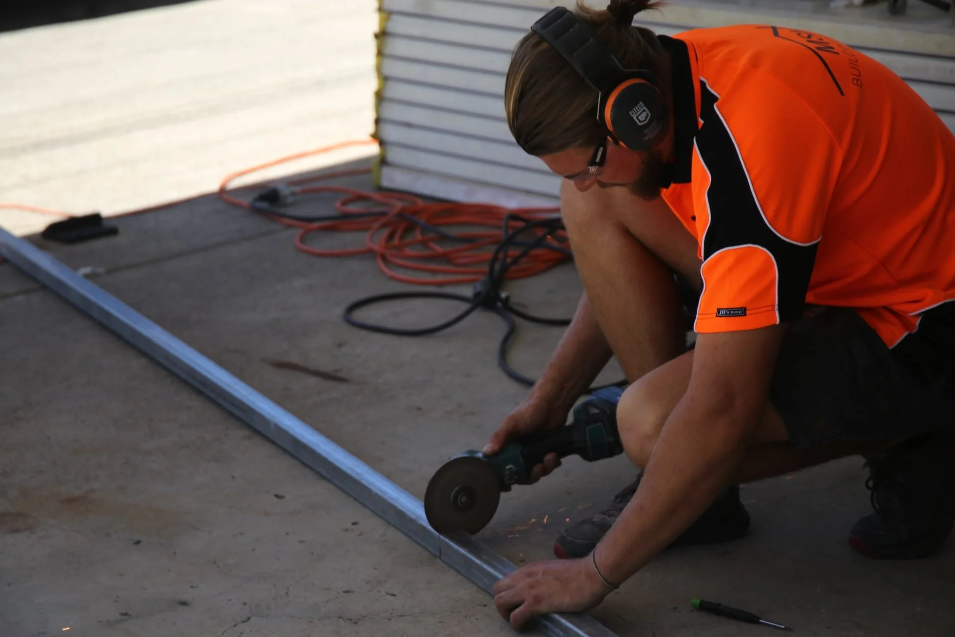 Person wearing orange and black shirt, safety glasses, and ear protection uses a handheld tool to cut a metal rod on a concrete surface. Cables and a metal sheet are visible in the background.