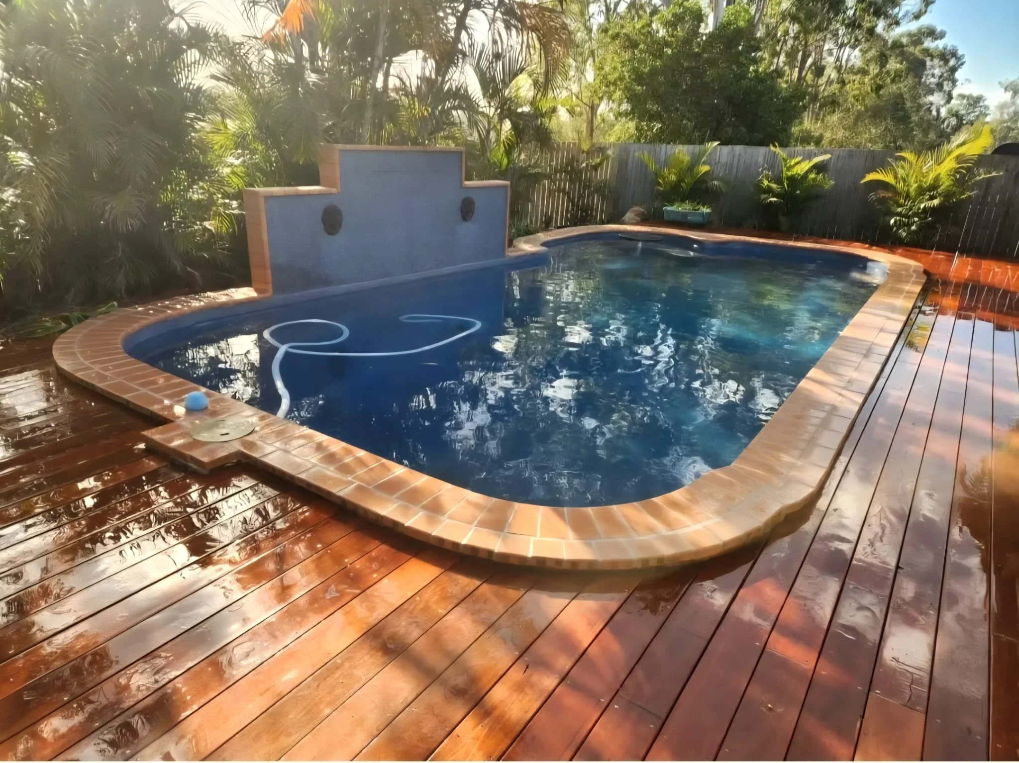 A backyard swimming pool with a wooden deck, surrounded by lush green plants and trees. The pool has a small fountain feature at one end and a blue tile interior. The scene is bright and sunny.