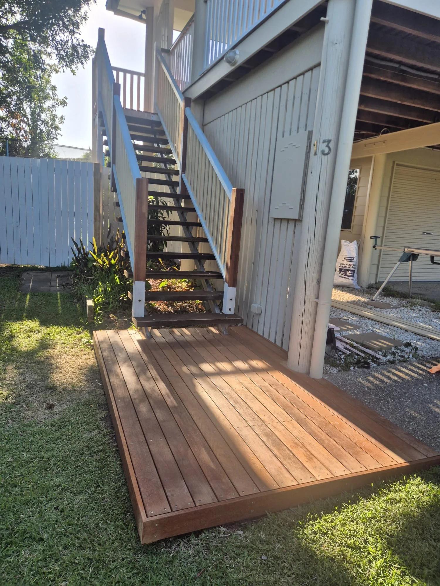 Wooden staircase with handrails leading to a second floor, placed on a wooden deck outside of a house with vertical siding and the number 13 visible. The area around the stairs has grass, plants, and a neighboring house with a garage door.