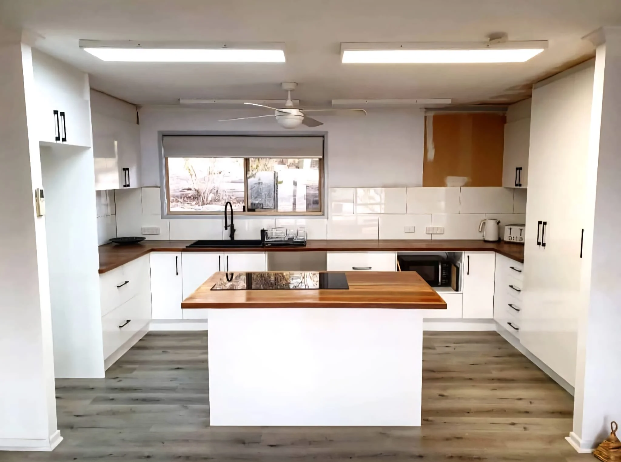 Empty kitchen with white cabinetry, wooden countertops, a kitchen island with a wooden top, a window behind the sink, and various appliances and utensils.