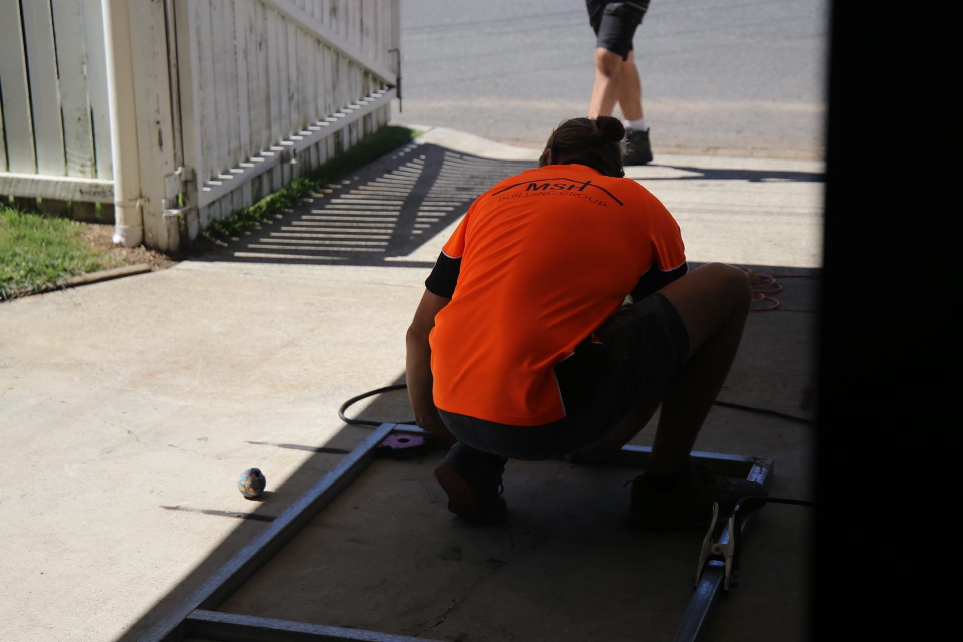 Person in orange shirt crouching on ground working with metal frame outdoors, shadowed area in the foreground, white fence and person walking in background.