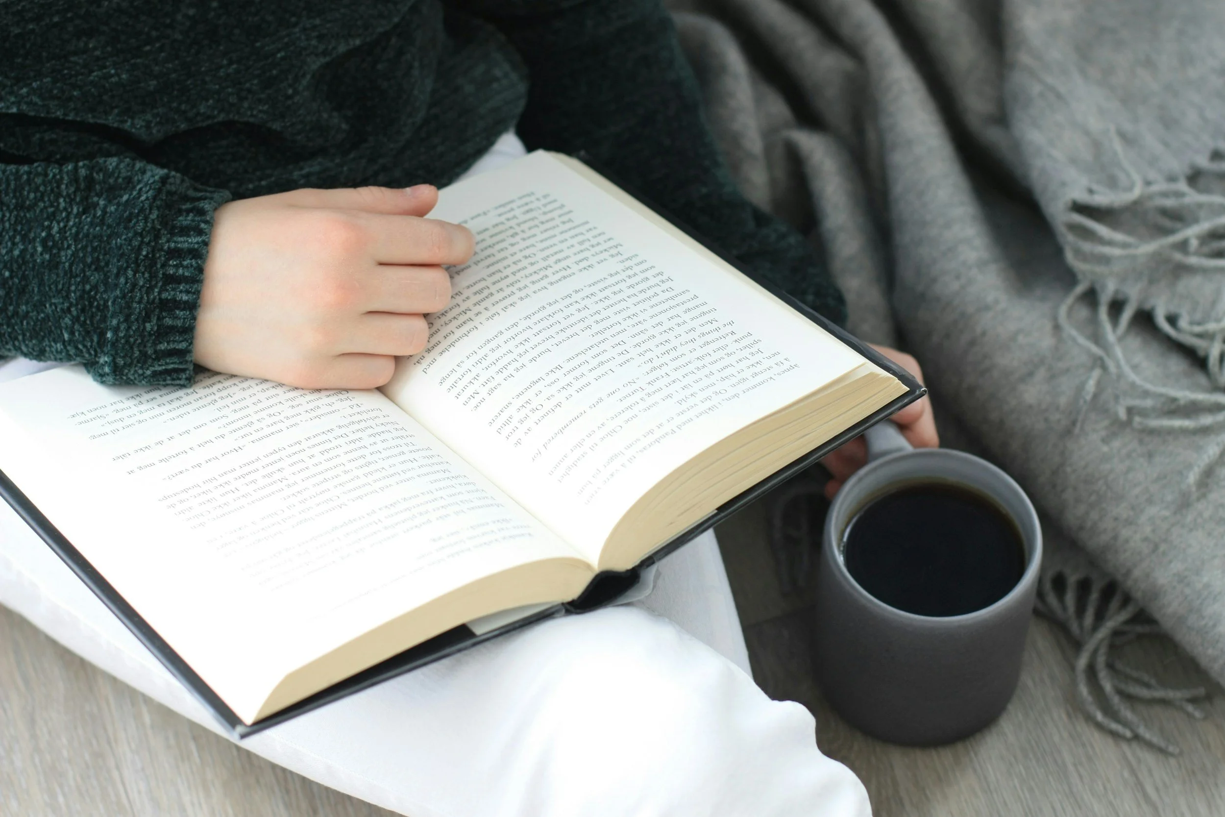Person reading a book with a cup of coffee nearby, sitting on a couch with gray blankets.