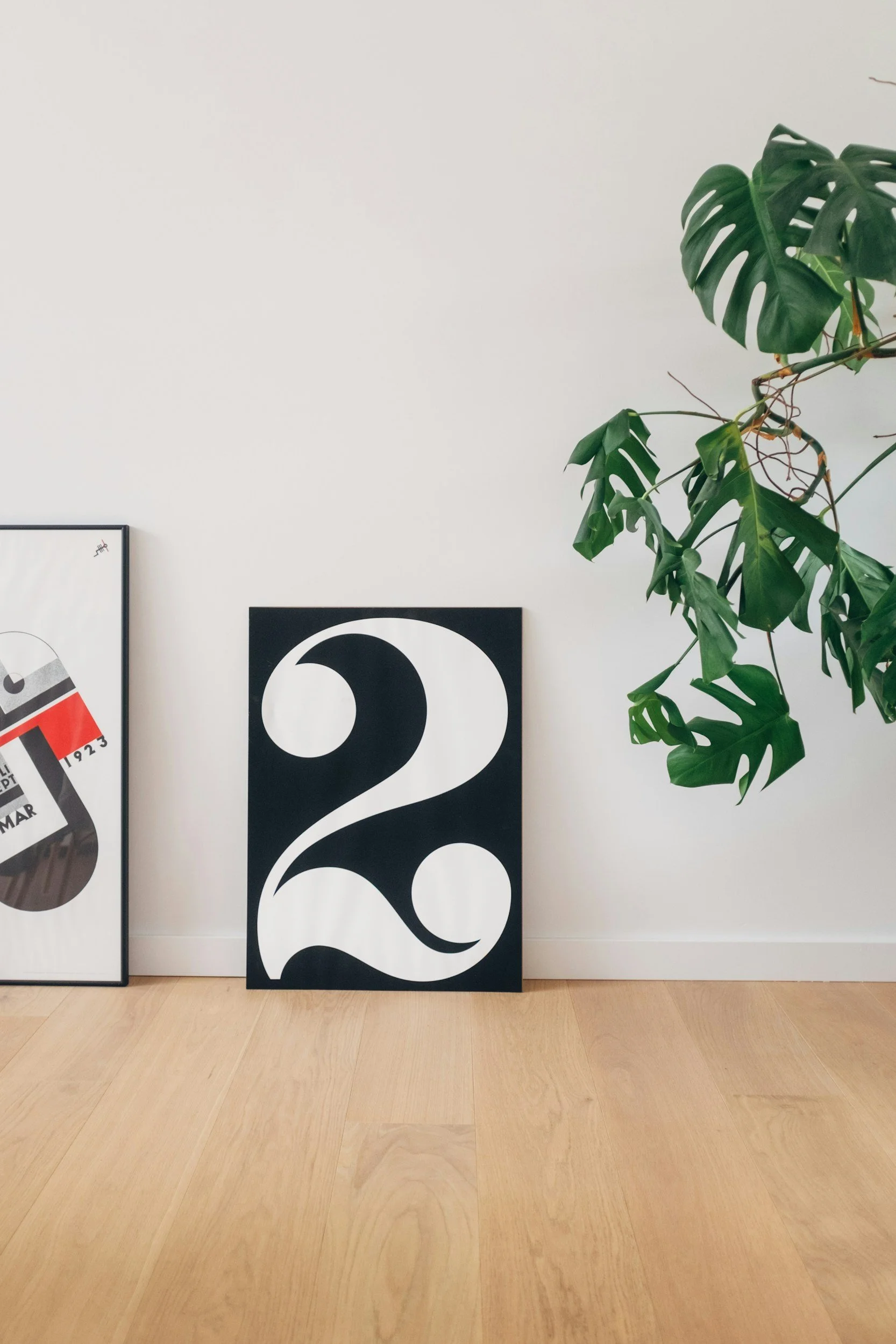 Black and white art print with a large number 2 leaning against a white wall, next to a potted plant with large green leaves.