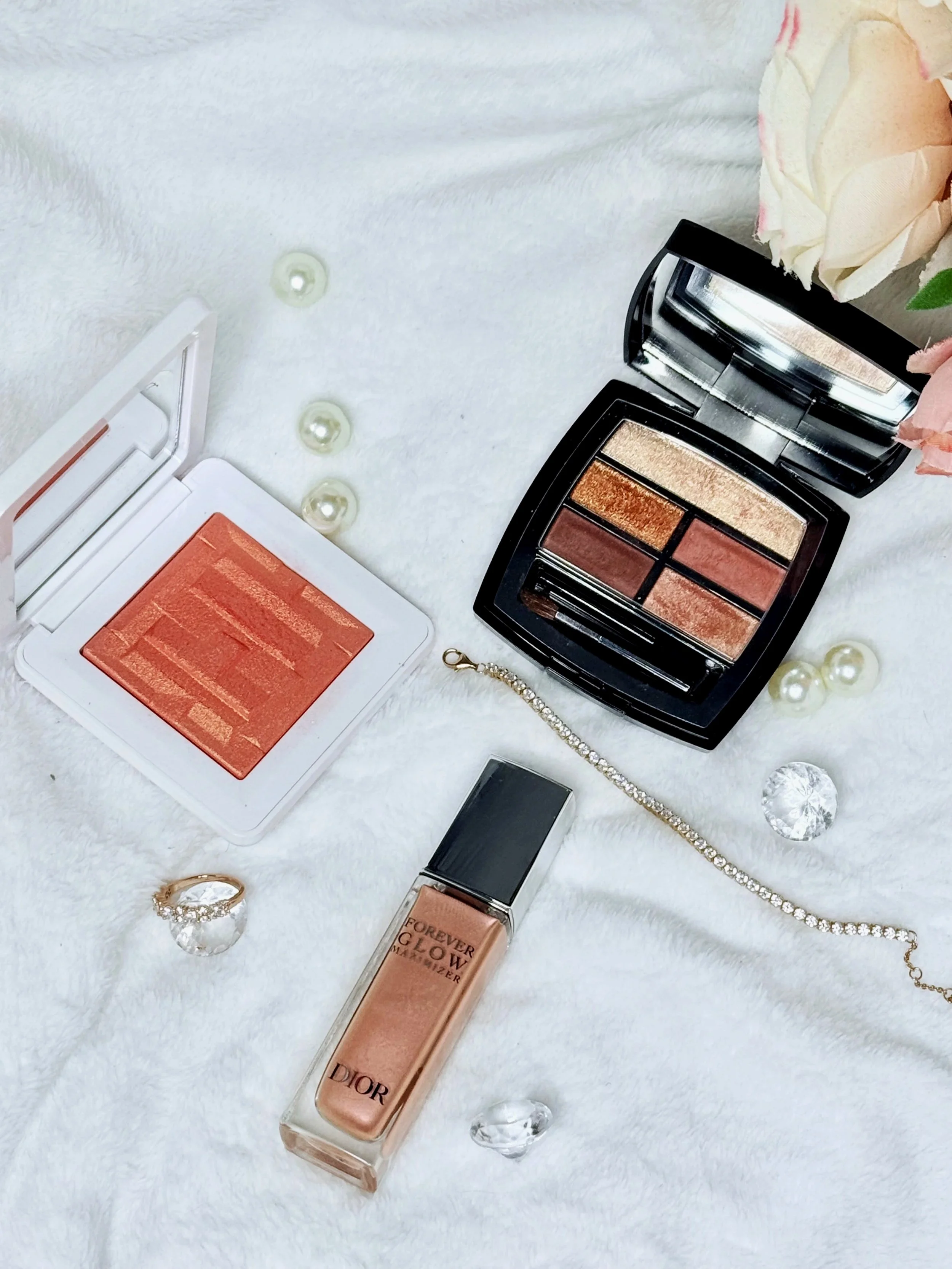 Open Dior Forever Glow foundation bottle, rectangular black palette with eyeshadows, peach blush, pearl necklace, jewelry, and decorative crystals on a white furry surface, topped with peach and white roses.