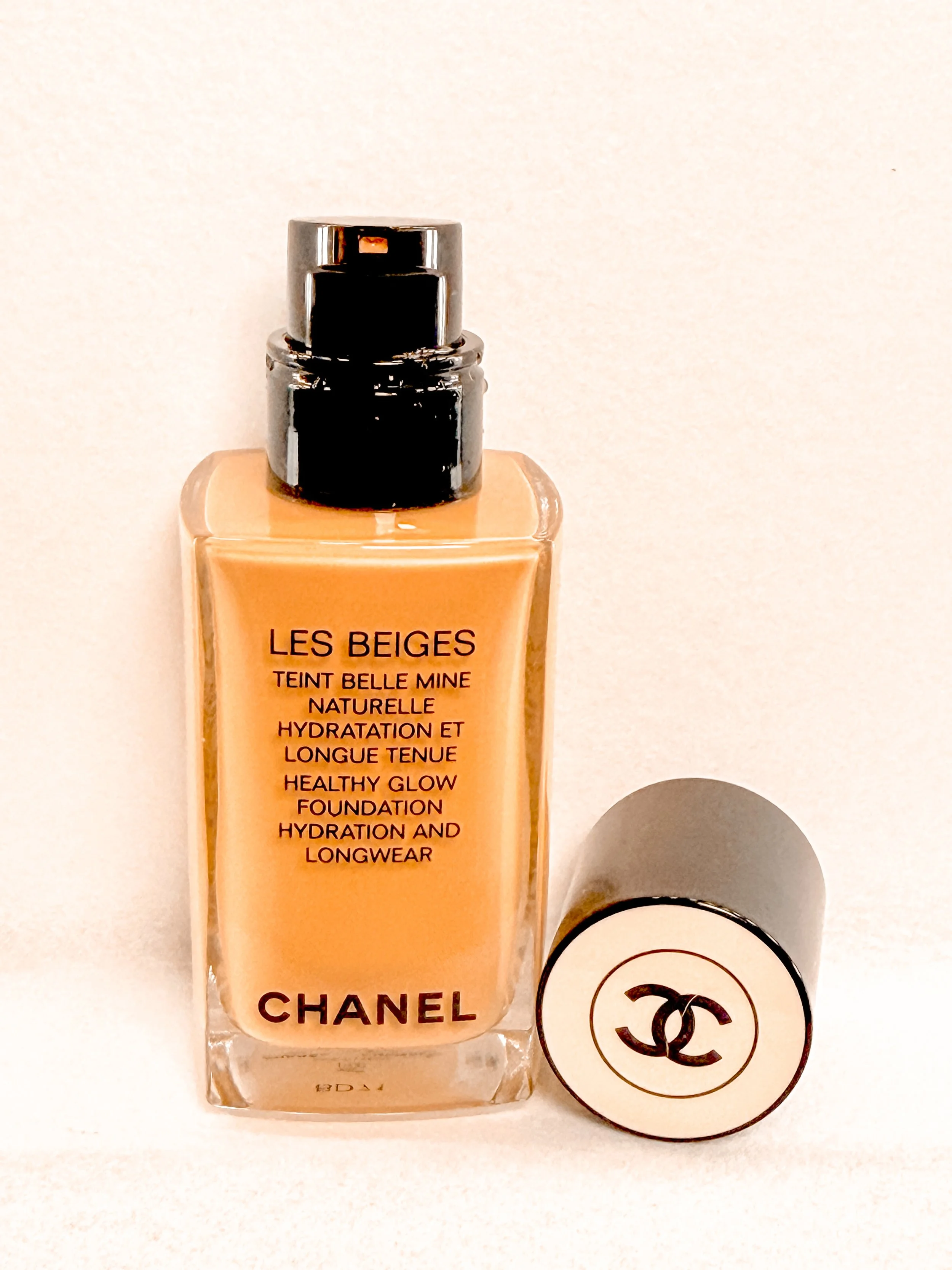 A beige-colored Chanel Les Beiges foundation bottle with a shiny black cap, placed on a white surface alongside a round makeup product with the Chanel logo.