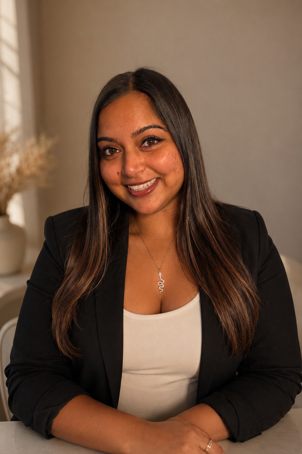 A woman with long dark hair, wearing a black blazer and a white top, smiling at the camera, with a necklace that has a snake pendant.