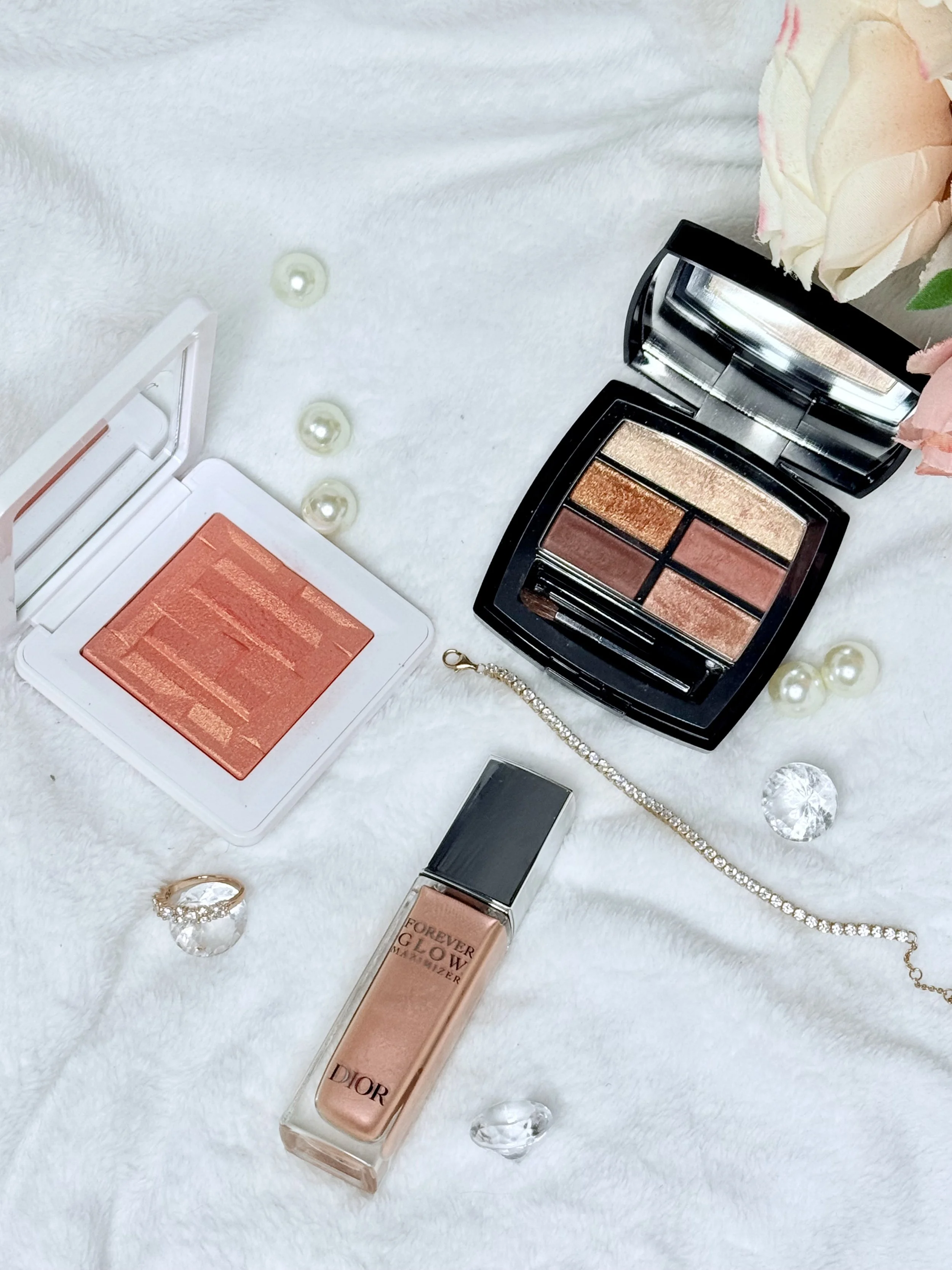 Makeup products including a peach-colored blush, a peachy eyeshadow palette with four shades, a bottle of Dior Forever Glow foundation, a black compact mirror, a pearl bracelet, and decorative pearls and crystals on a white, fluffy surface.