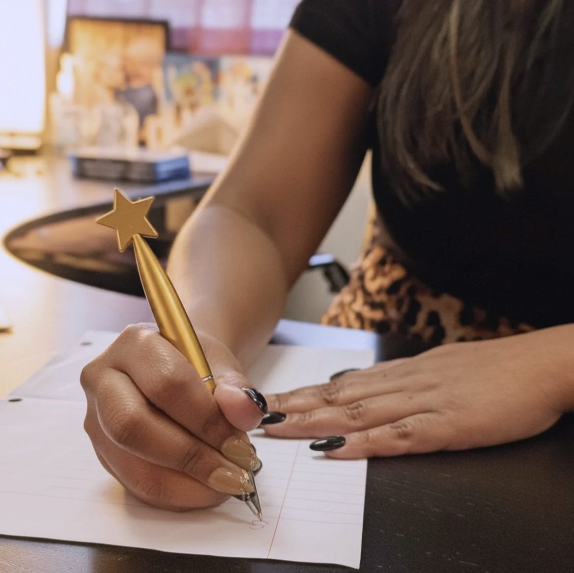 Person writing in a notebook with a gold star-shaped pencil, sitting at a desk in an office or classroom setting.