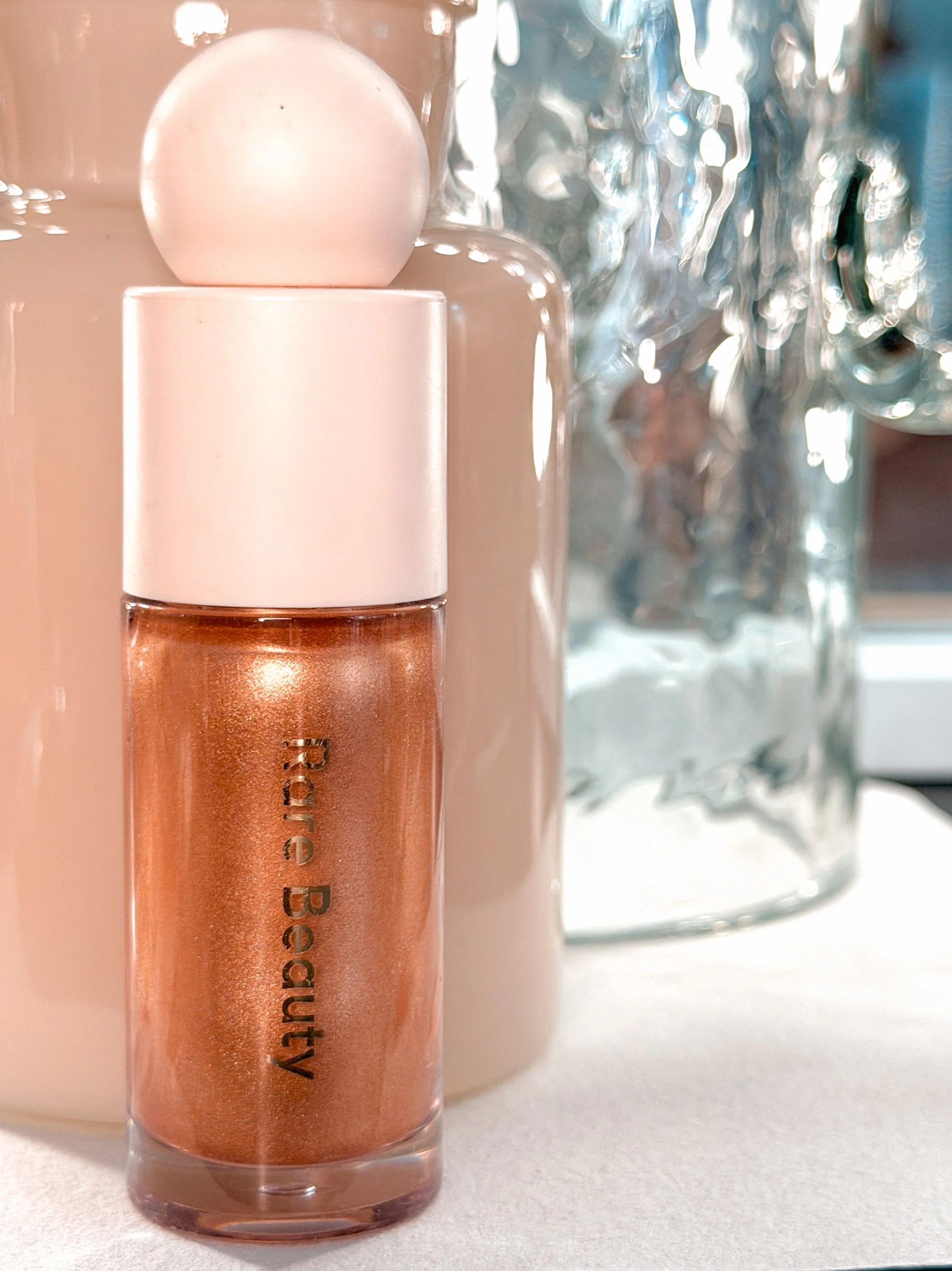 Close-up of a lip gloss or liquid lipstick tube with a creamy, peachy hue, with the text 'Anytime Be...' visible, placed on a light-colored surface with a blurred crystal-like object in the background.