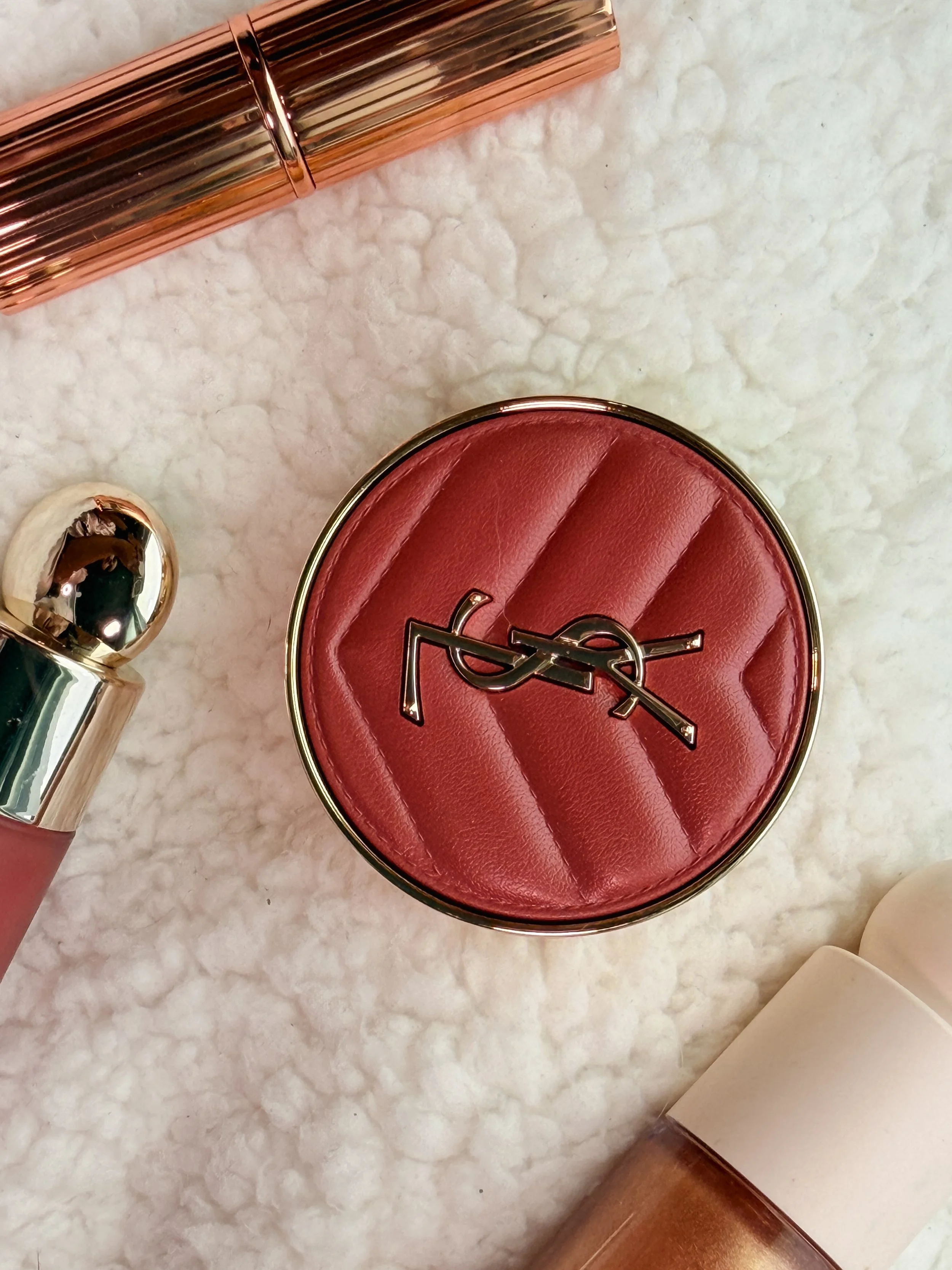 Close-up of a red Yves Saint Laurent compact on a marble surface, with part of a rose gold makeup brush and a white object visible.