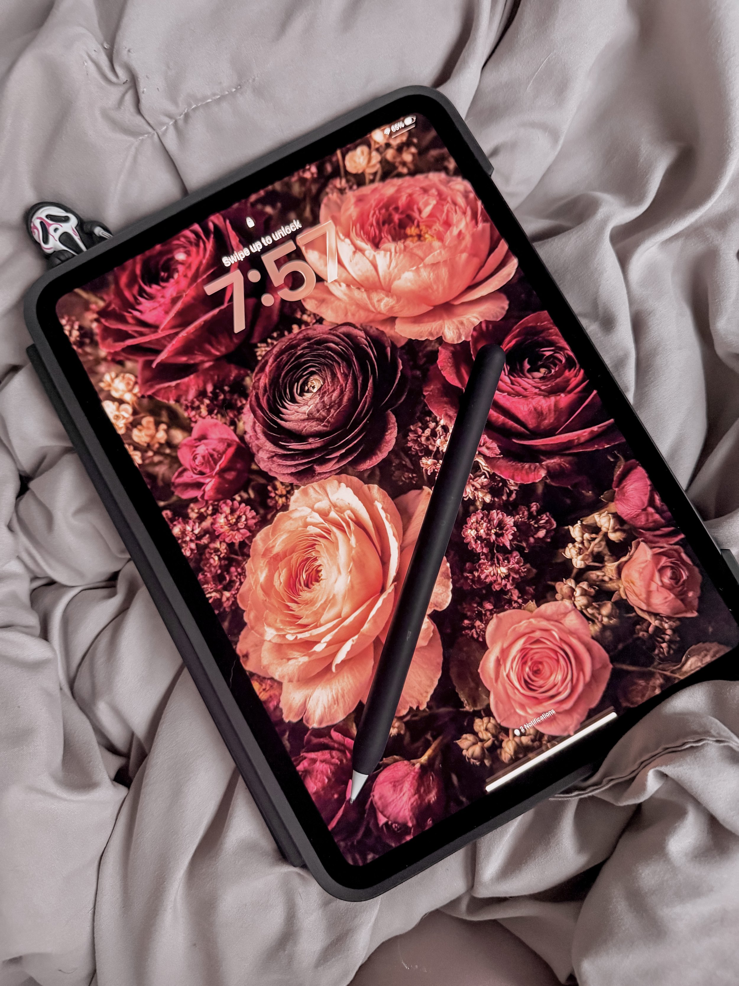 A tablet with a floral wallpaper of pink and purple flowers, showing the time 7:57, resting on a crumpled gray bedsheet with a stylus on the screen.