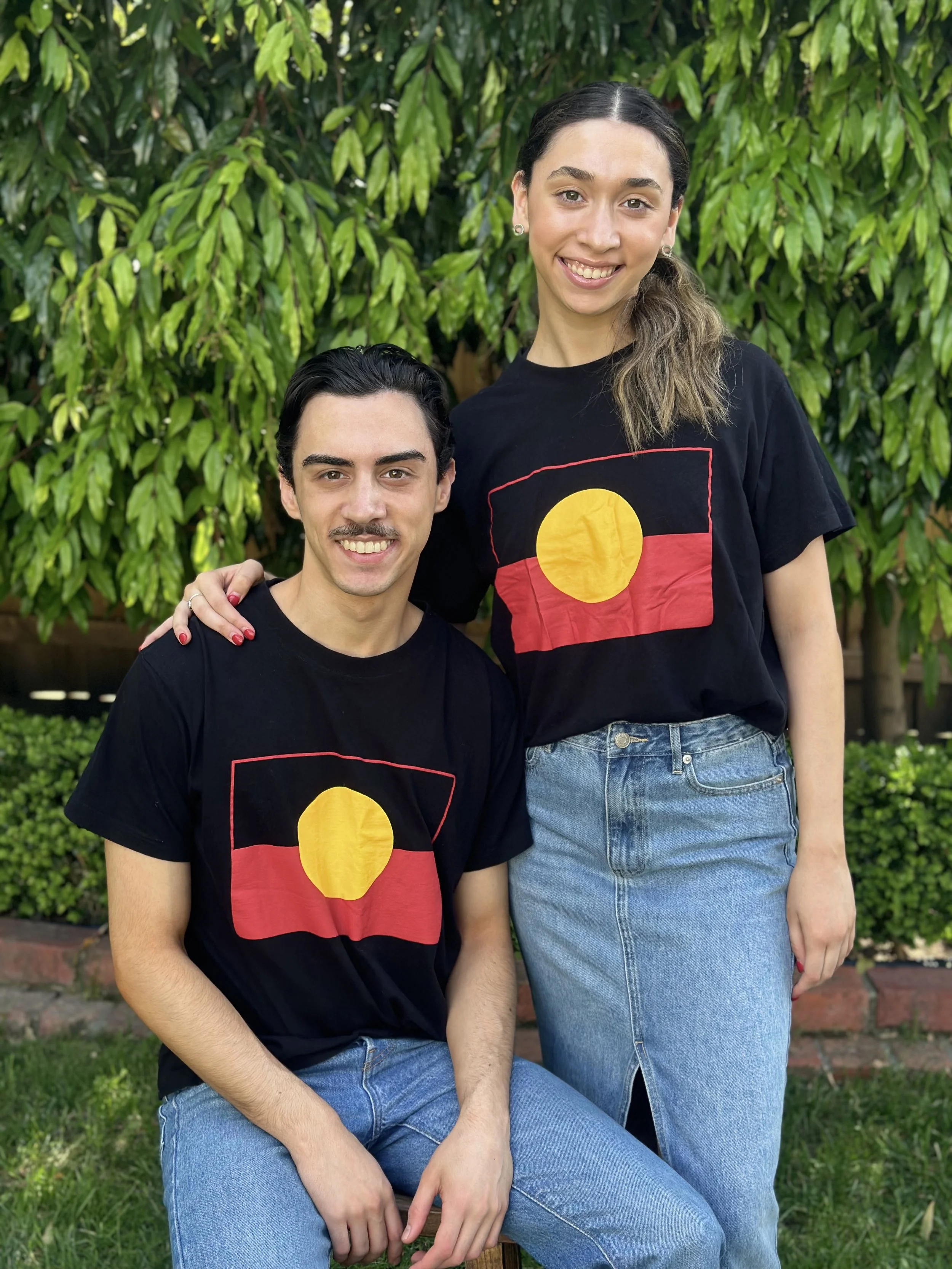 Two people outside, wearing black t-shirts with an Aboriginal Australian flag design. One person is sitting, and the other is standing, with greenery in the background.
