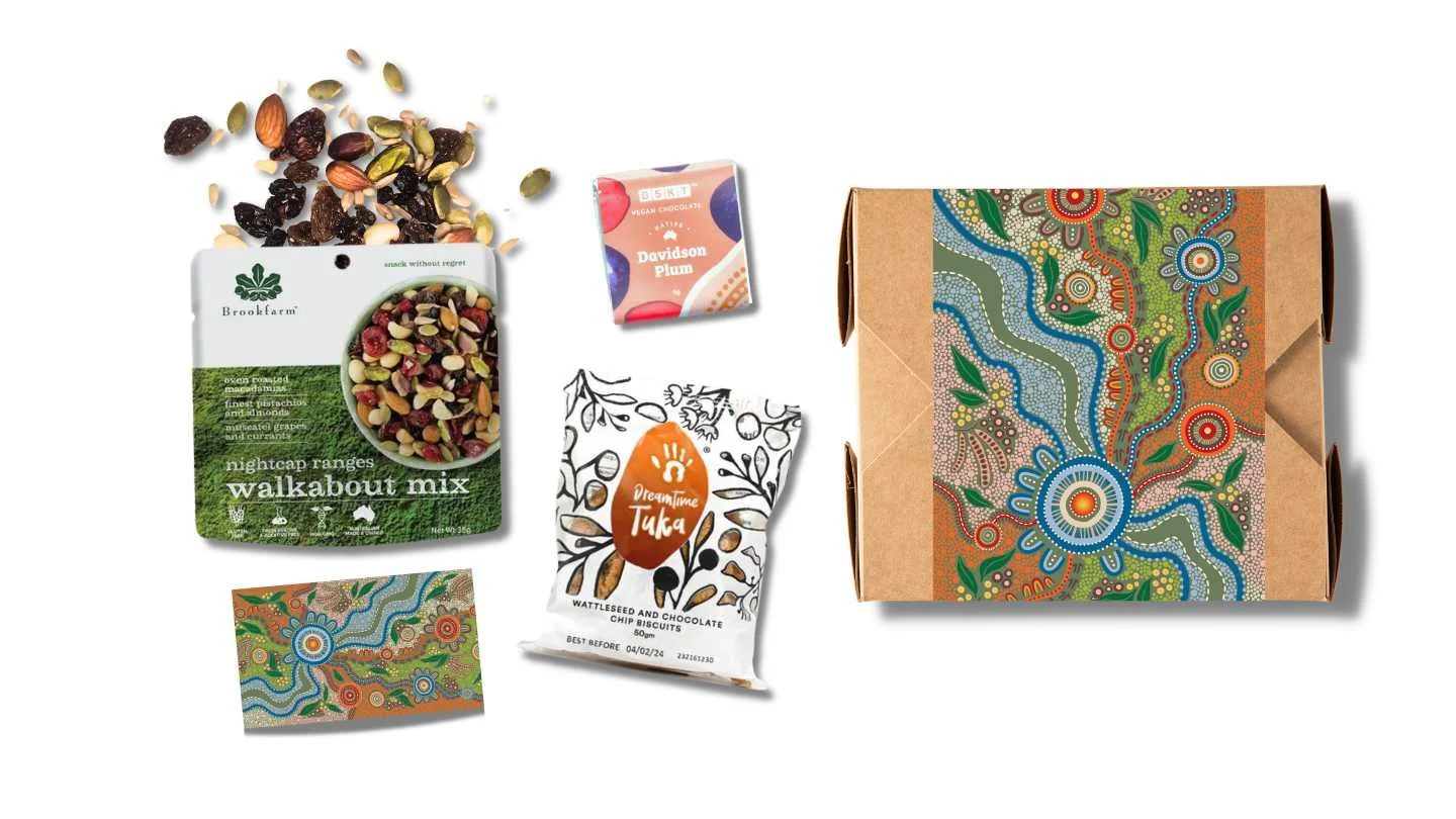 Assorted snack packages including Brookfarm's 'Walkabout Mix', BSKT's vegan chocolate with Davidson Plum, Dreamtime Tuka's wattleseed and chocolate chip biscuits, and a decorative box with Aboriginal-style patterns.