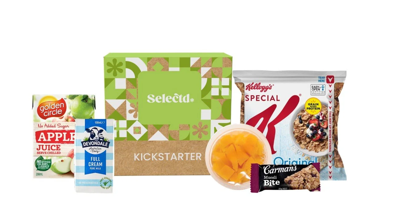 A selection of food and beverage items including apple juice, full cream milk, a bowl of diced mango, a cereal bar, a bag of Special K cereal, a Kickstarter box, and a packet of Carman's muesli bites.