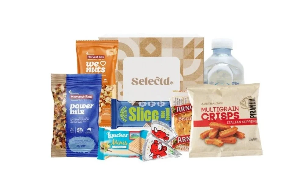 Assorted snack items and bottled water in front of a Selecta box. Items include Harvest Box power mix, We ♥ nuts, Loacker Minis, Slice snack, Arnott's biscuits, multigrain crisps, and a small bottled water.