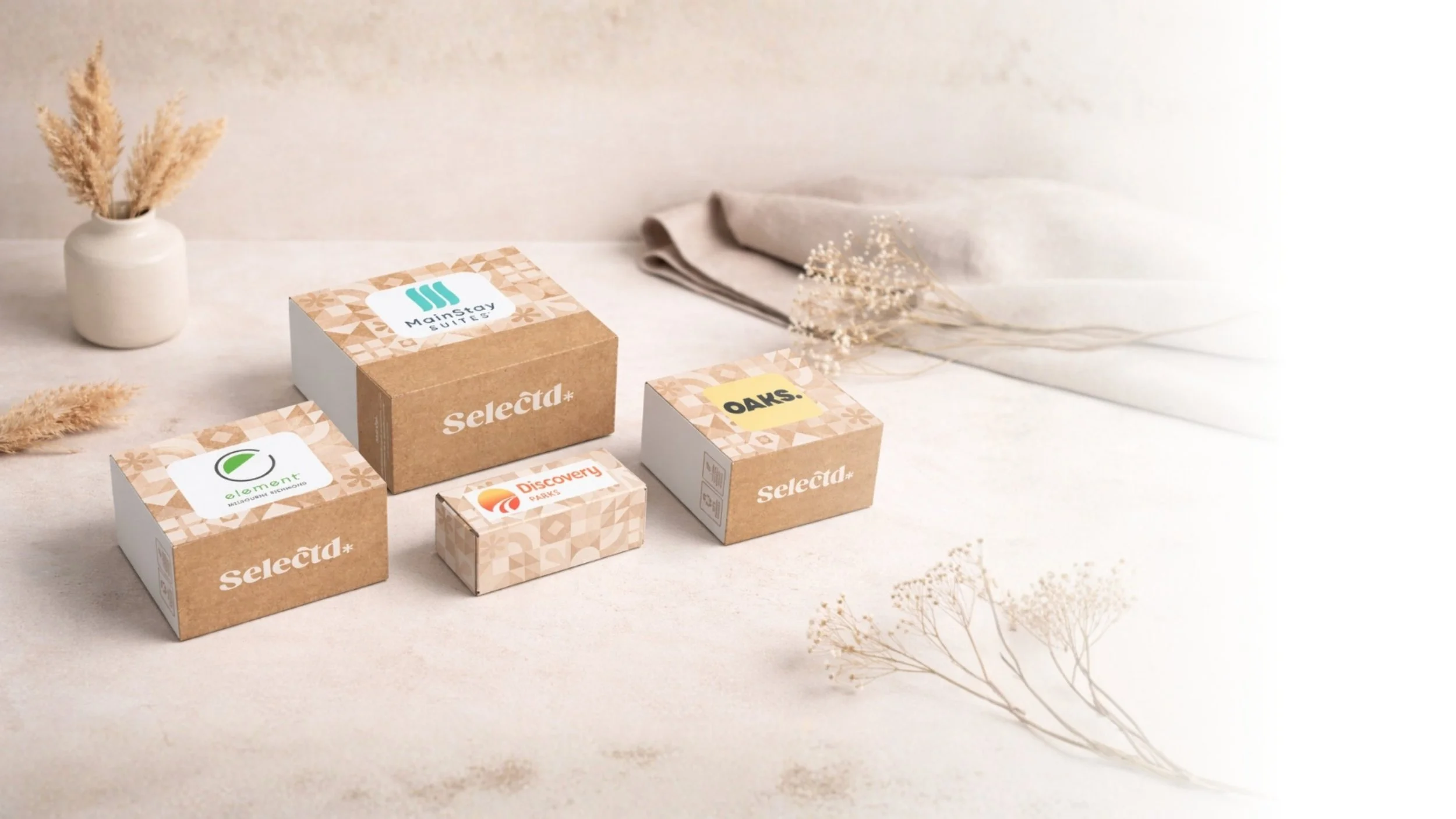 Several small and medium-sized boxes of assorted brands, a white vase with beige dried grasses, a folded light-colored cloth, and some dried flowers on a light surface.