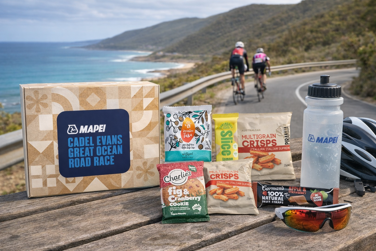 A collection of snacks, a water bottle, and cycling gear on a wooden surface by a coastal road with two cyclists riding in the background.