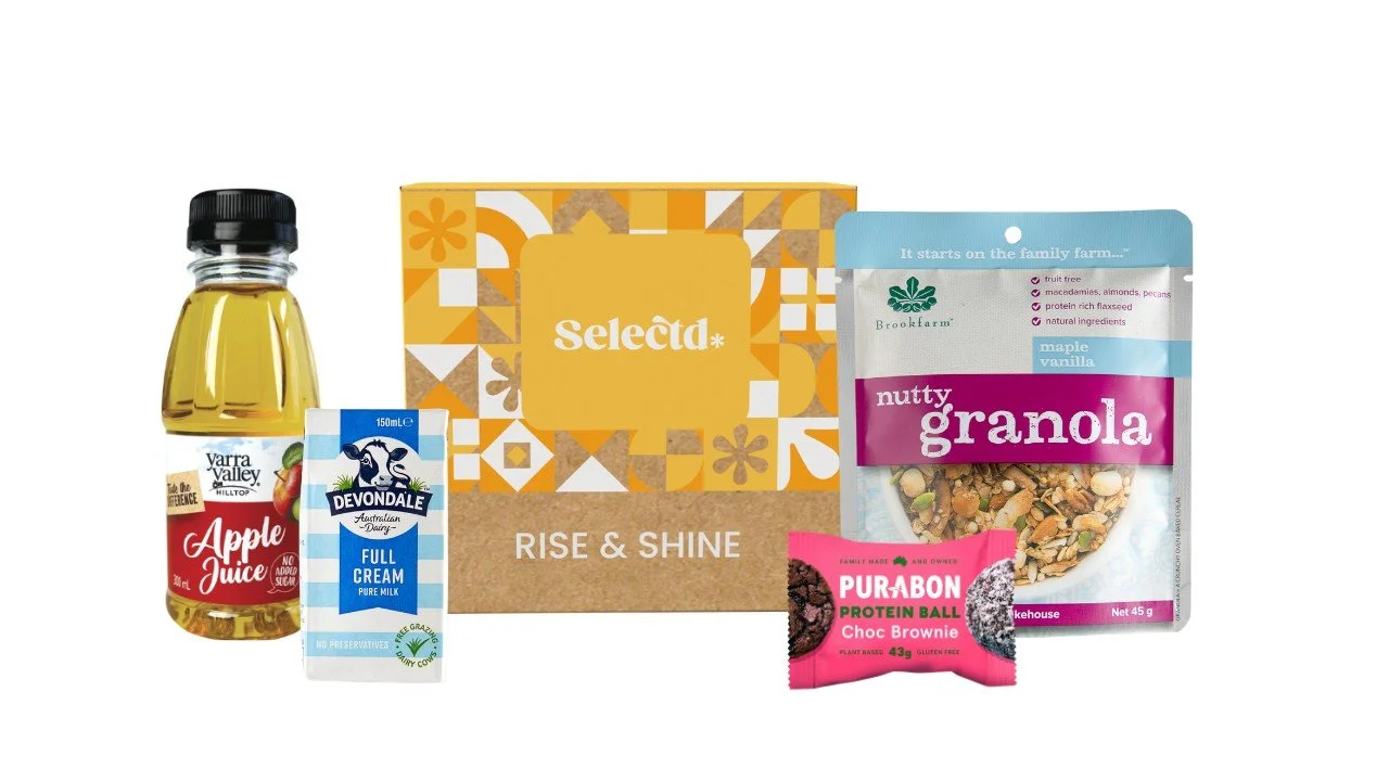 A variety of breakfast items including apple juice, full cream milk, a box labeled Selecta Rise & Shine, a bag of nutty granola, a Protein Ball Choc Brownie snack, and a small package of Purabon protein bites.